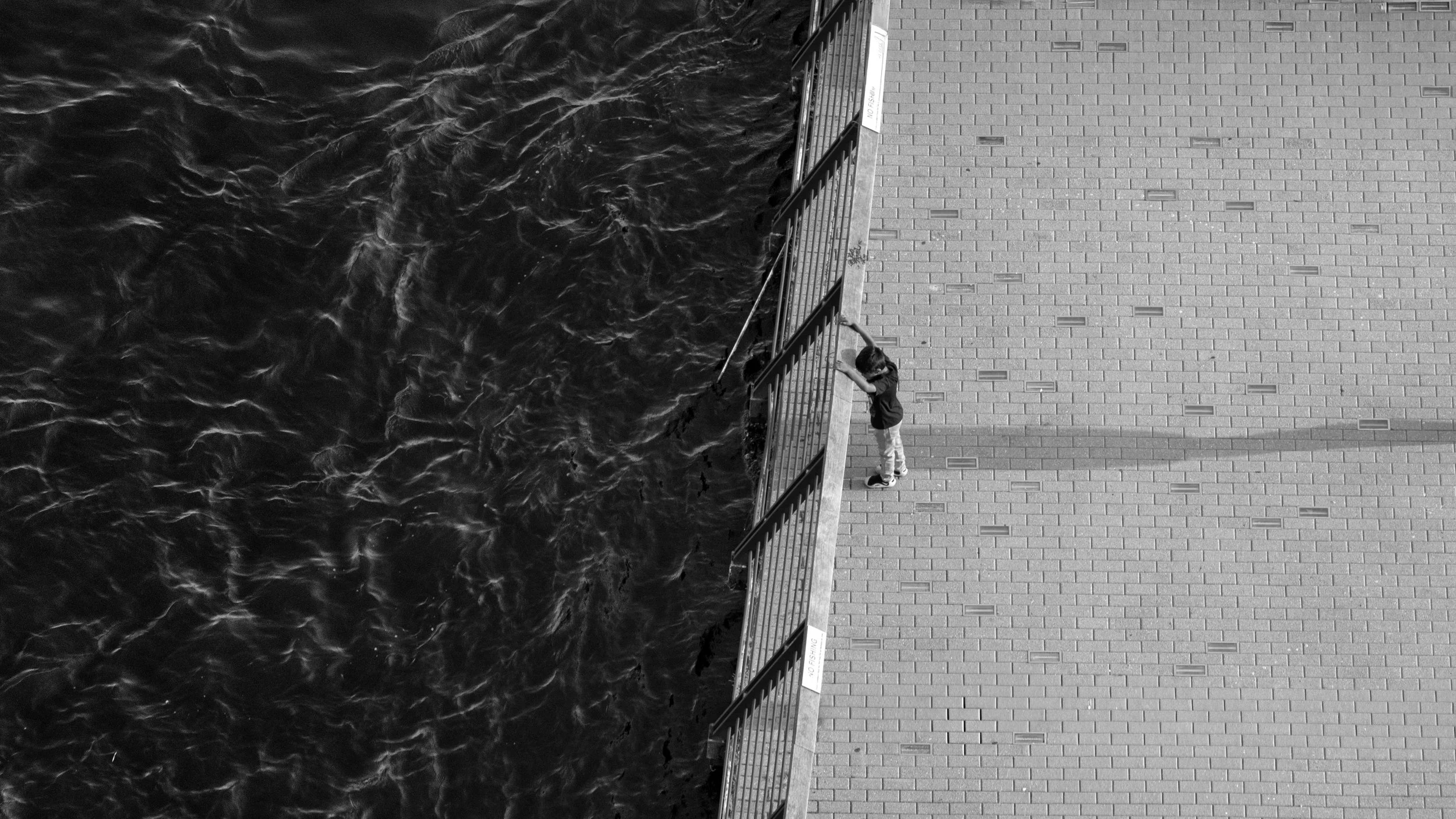 A child standing on a paved area near a metal railing, looking out over water, in black and white.