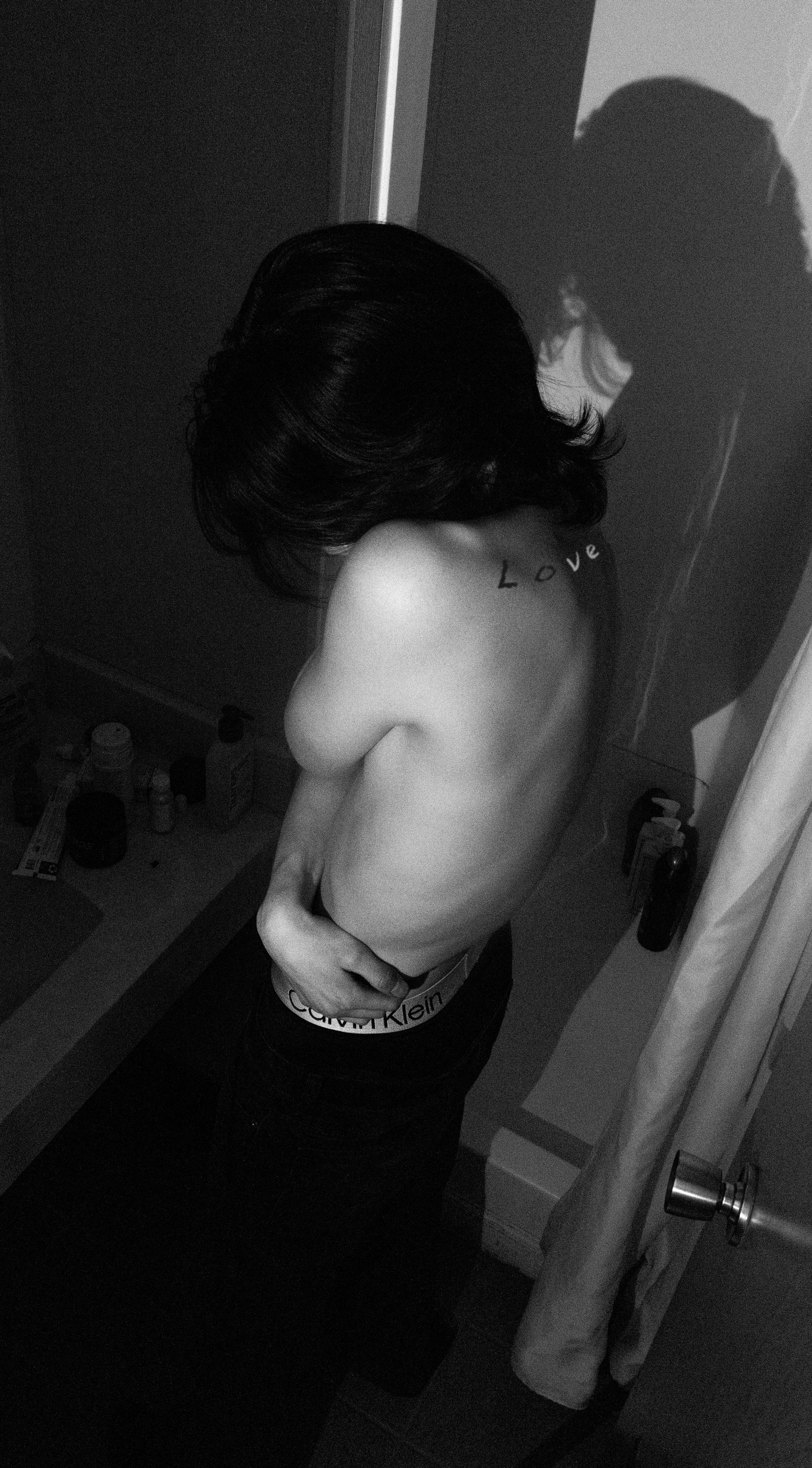 A woman with dark hair standing in a bathroom, wearing black Calvin Klein underwear with the waistband visible, her left arm covering her chest, a tattoo reading 'Love' on her shoulder blade, and her shadow cast on the wall behind her.