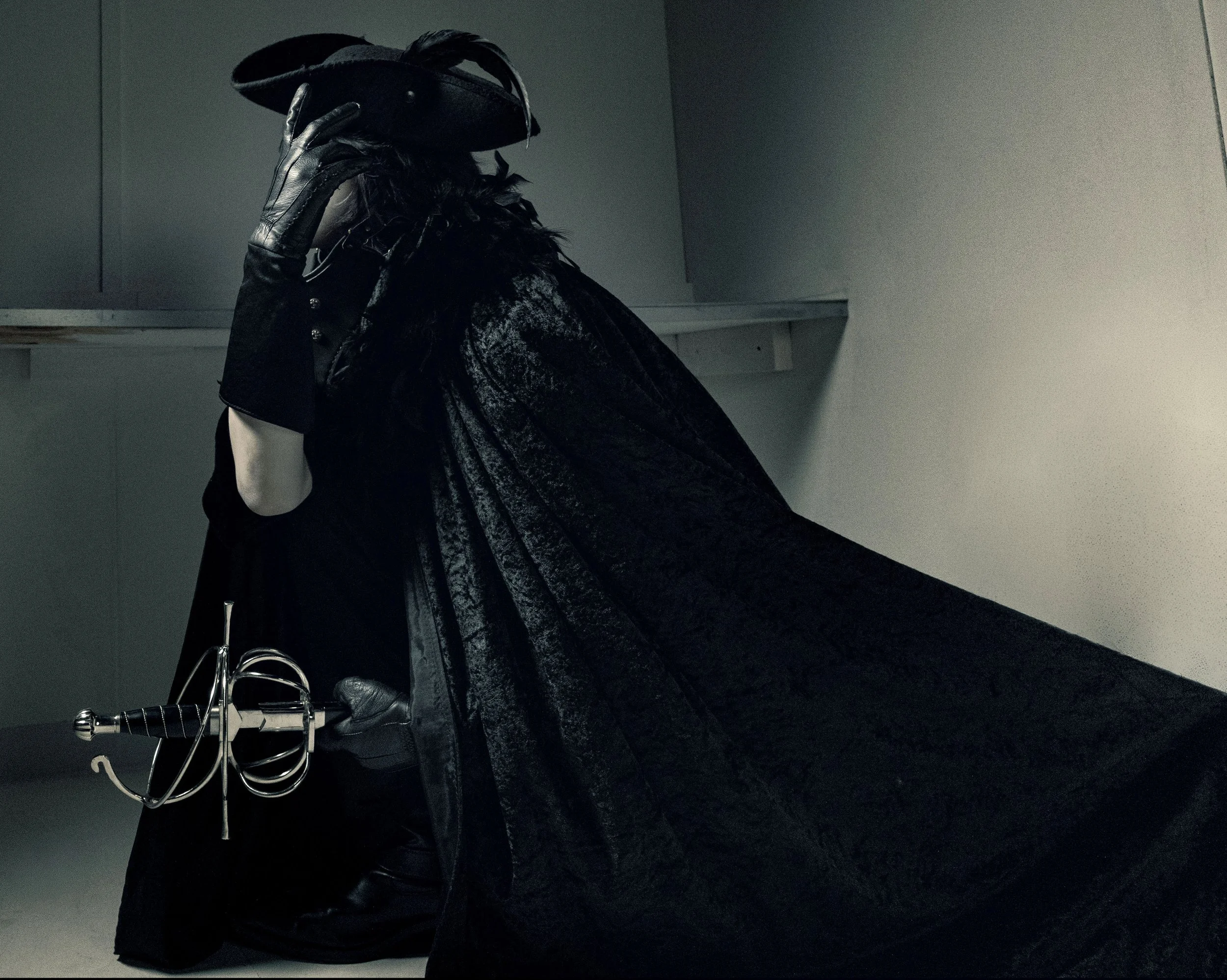 A person dressed in black wearing leather gloves and a feathered cape, holding a hat in front of their face while standing in front of a dark wall. They are also holding a fencing sword.
