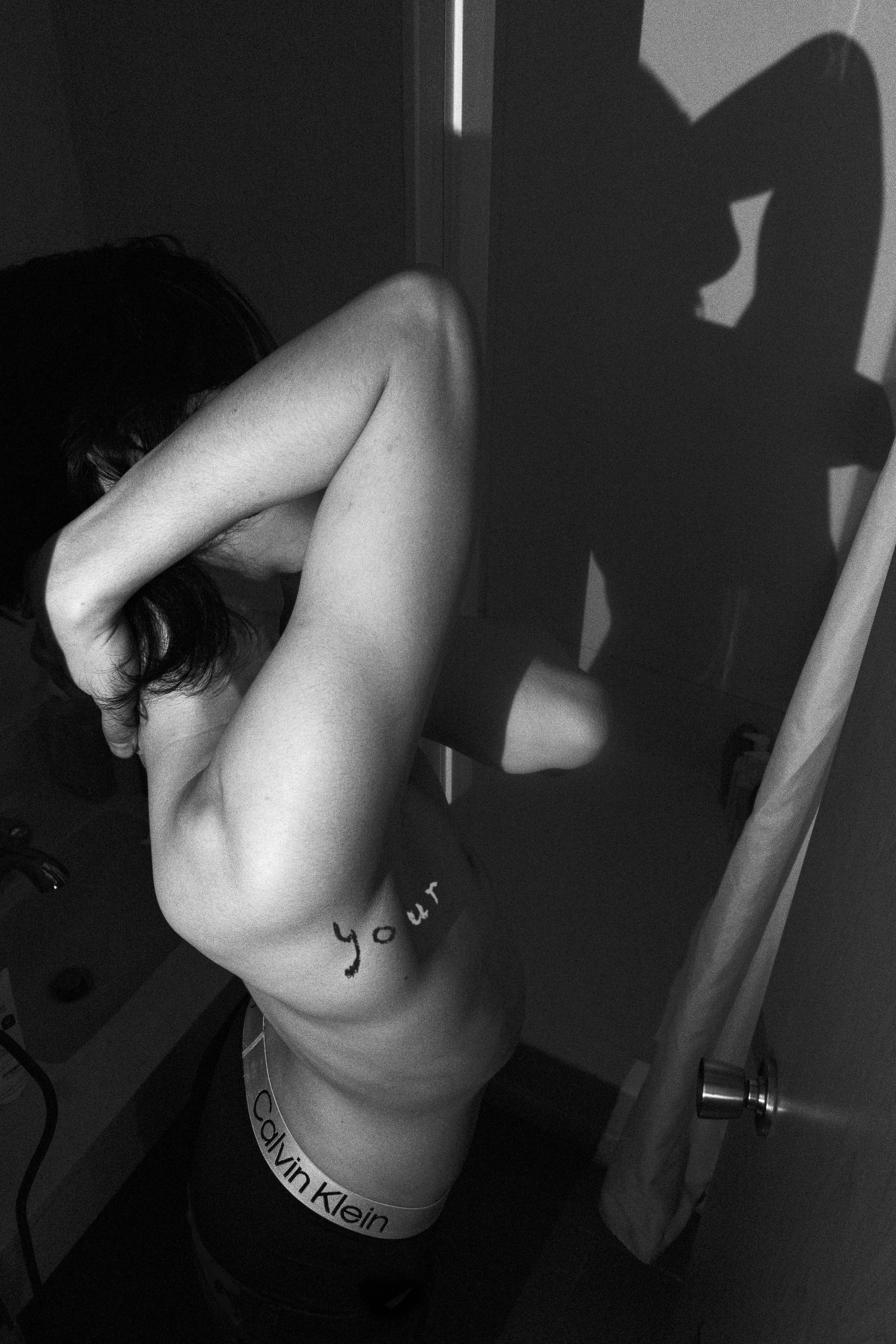 A person is taking a mirror selfie in a bathroom, wearing Calvin Klein underwear, with their head bowed down and arm partially covering their face. Their shadow appears on the wall behind them.