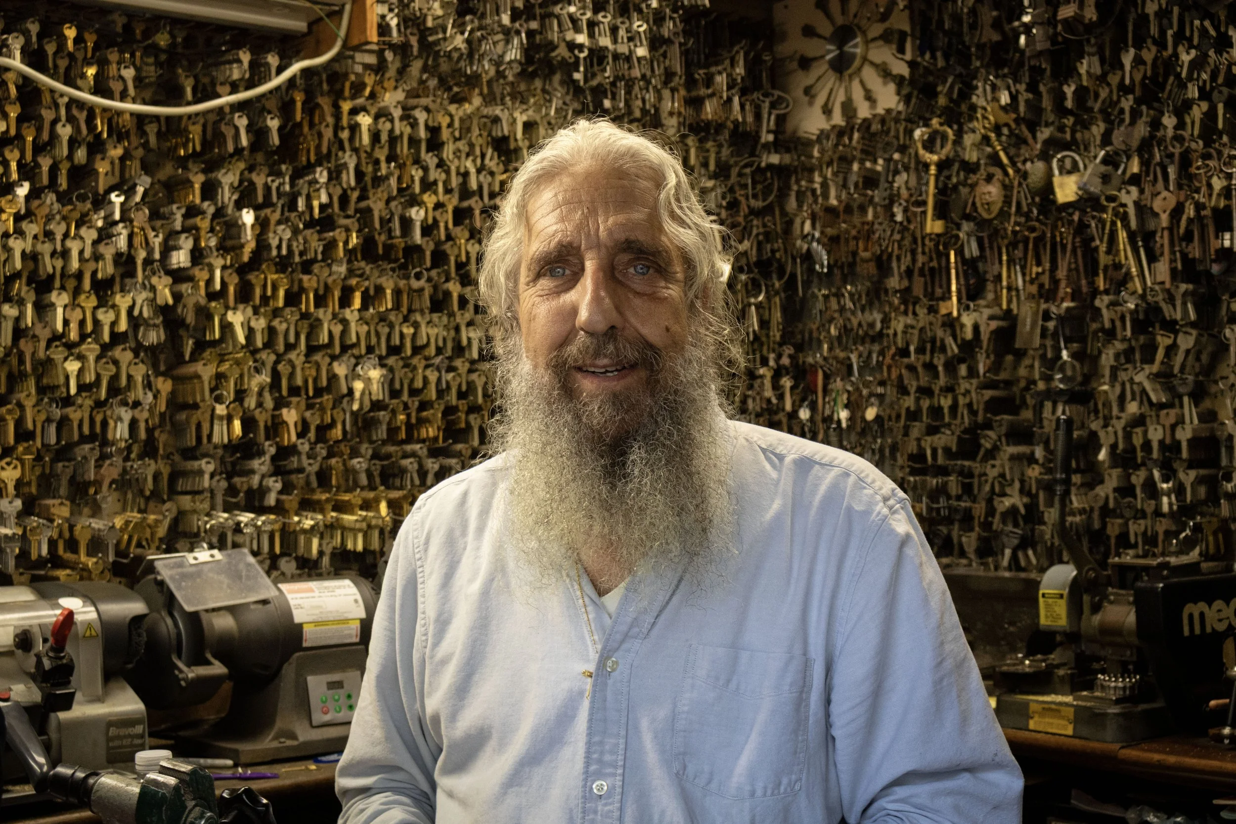 An older man with white hair and a beard wearing a white shirt standing in front of a large wall filled with hanging keys.