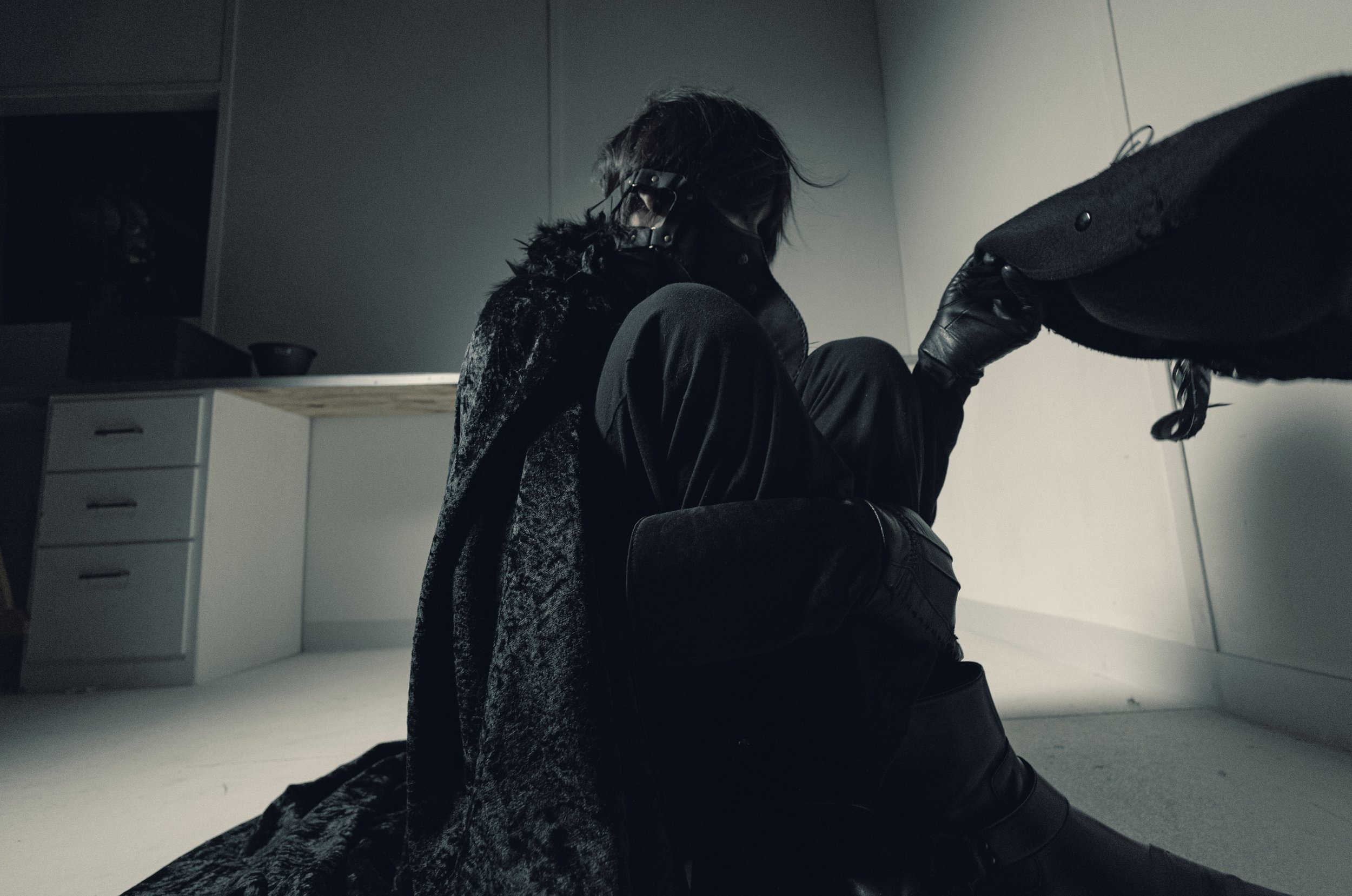 Person with dark hair, wearing black gloves and a fur-lined coat, sitting on the floor in a dimly lit room with white cabinets.