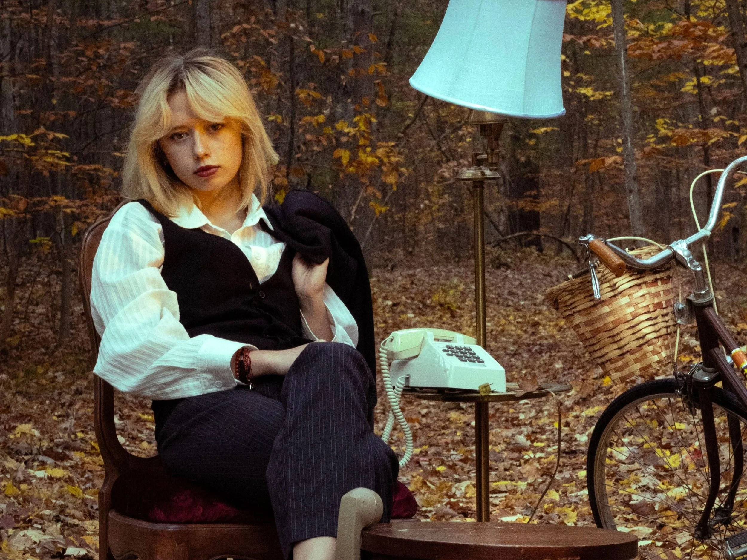 A young woman with blonde hair sitting outdoors in a forest with autumn leaves on the ground, next to a basket bike, a table with a landline phone, and a lamp. She is wearing a white shirt, a black vest, and dark pants, looking at the camera with a serious expression.