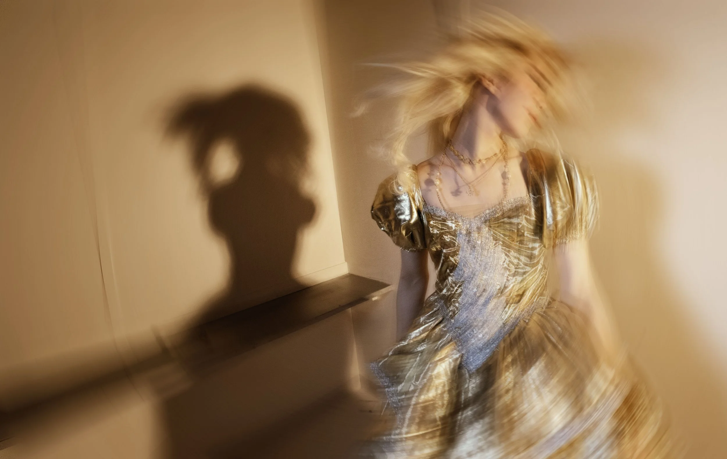 Blonde woman in a shiny, metallic gold dress with layered necklaces, twirling and blurry in motion, casting a shadow on the wall.