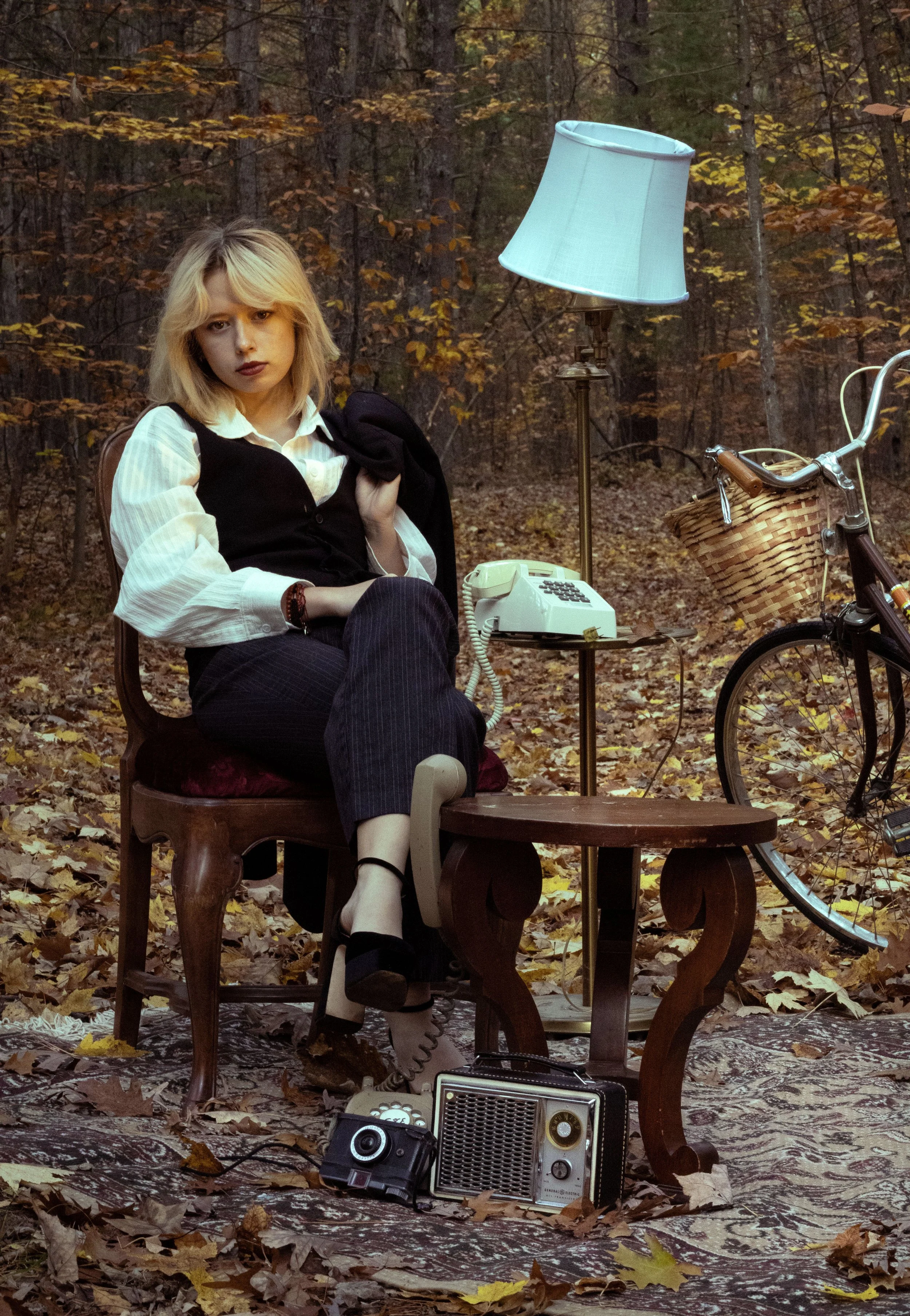 A woman with blonde hair sits on a wooden bench in an autumn forest, with fallen leaves on the ground. She wears a white blouse, black vest, and pinstripe pants, holding a black jacket over her shoulder. There is a vintage rotary phone on a small tab
