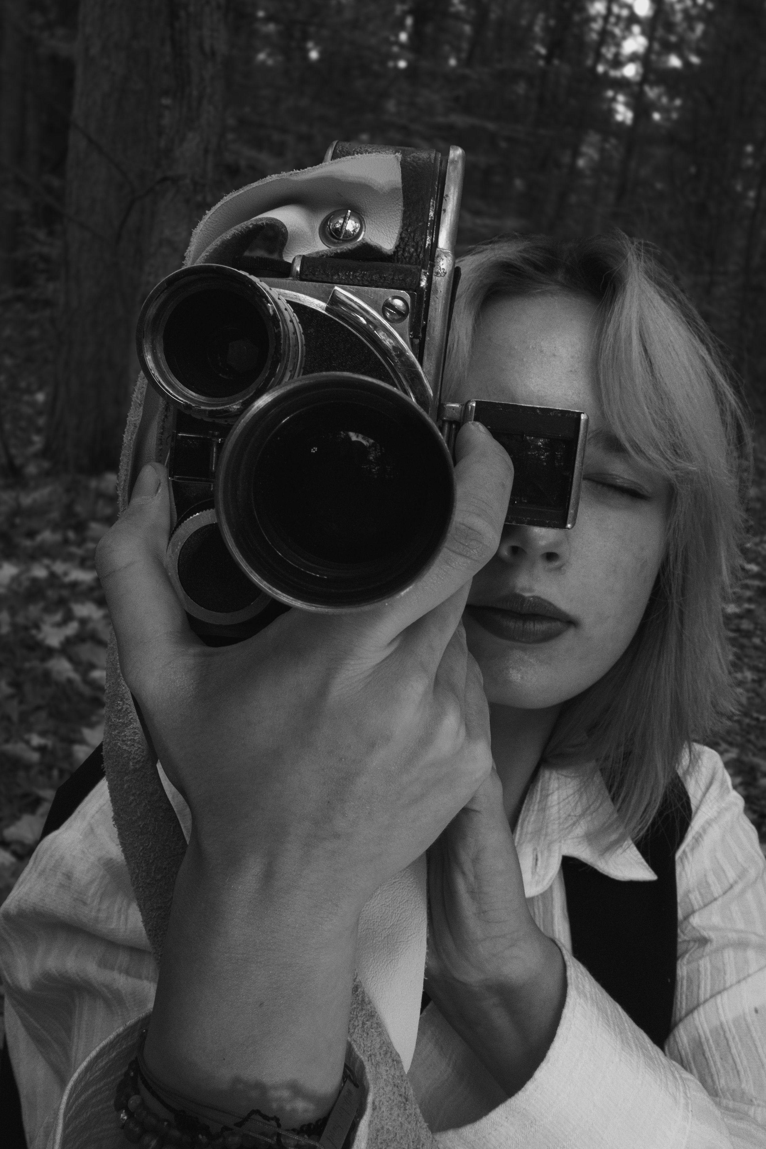 A woman is taking a selfie with a vintage camera in the woods, with trees and leaves in the background.