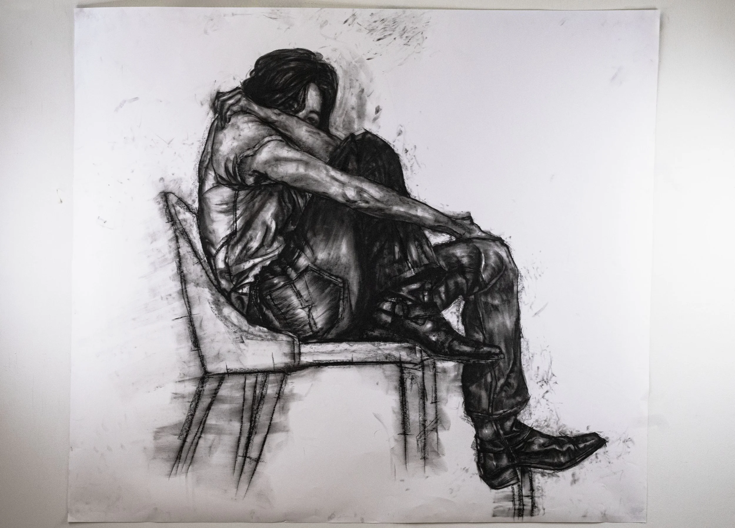 A black and white sketch of a person sitting with their knees pulled to their chest and their head resting on their arms, looking downward. The figure appears contemplative or sad.