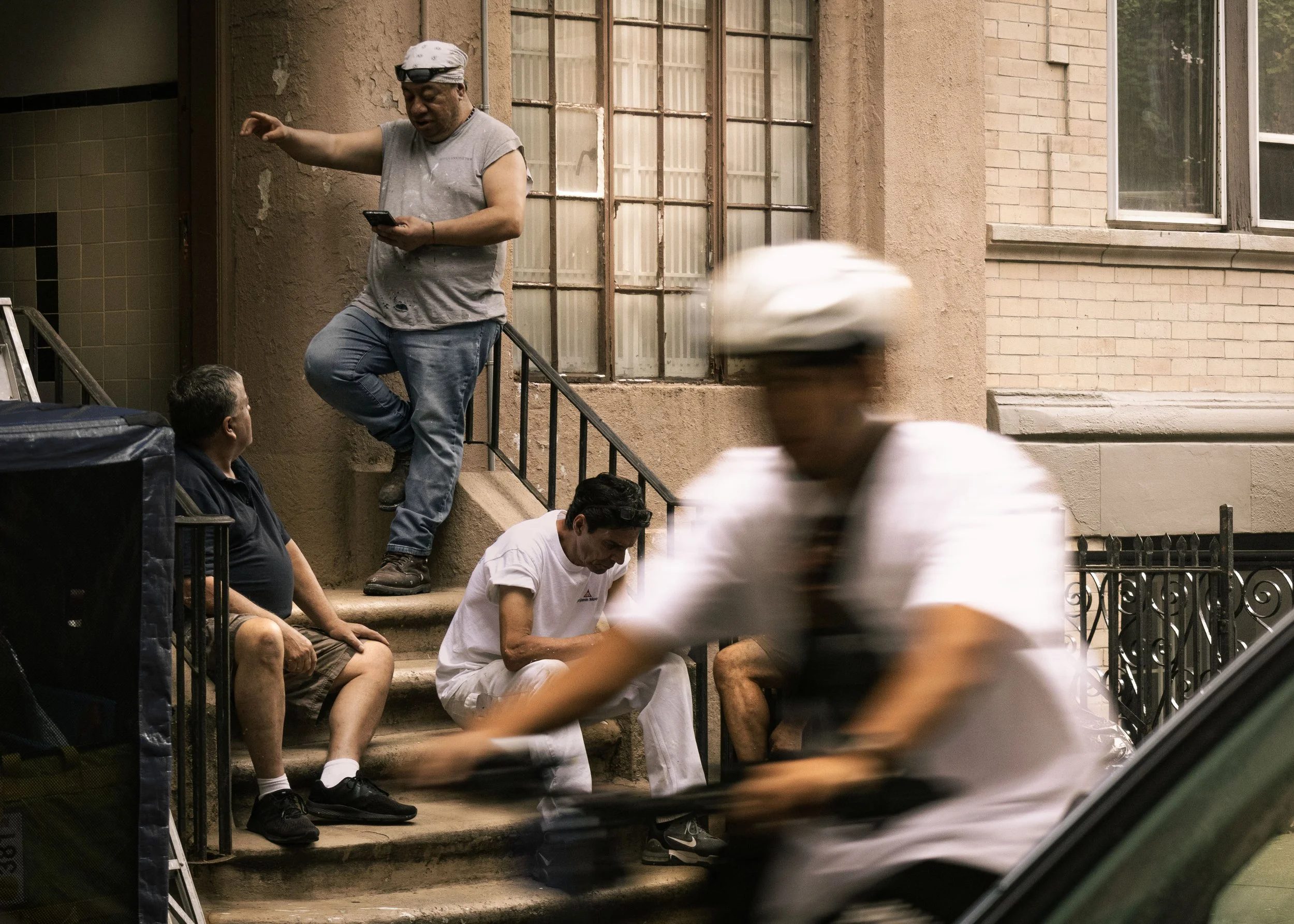 A man with a grey shirt and a cloth wrapping his head stands on a staircase, looking at his phone. Several people are sitting on the stairs and adult at the bottom is dressed in white, looking down. Blurred figures walk past in front of the camera.