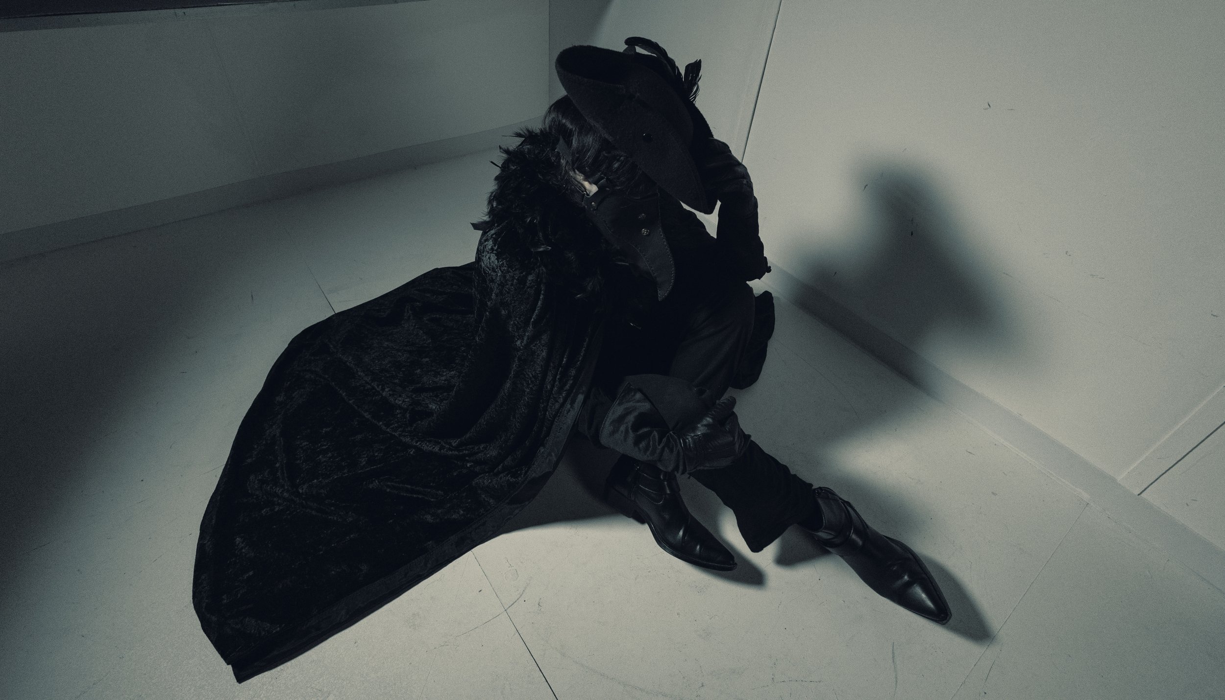 Person dressed in dark clothing sitting on the floor in a corner with their head down.