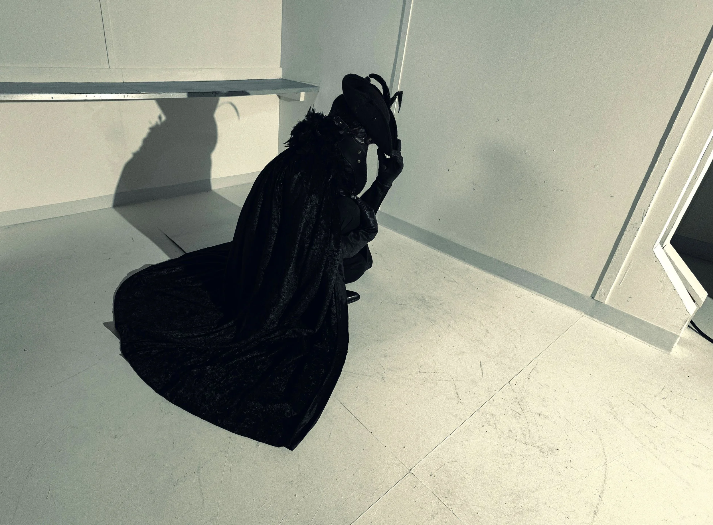 A person dressed in black, wearing gloves, and a hat, kneeling on the floor and adjusting something on their head, in a white room with minimal furniture.