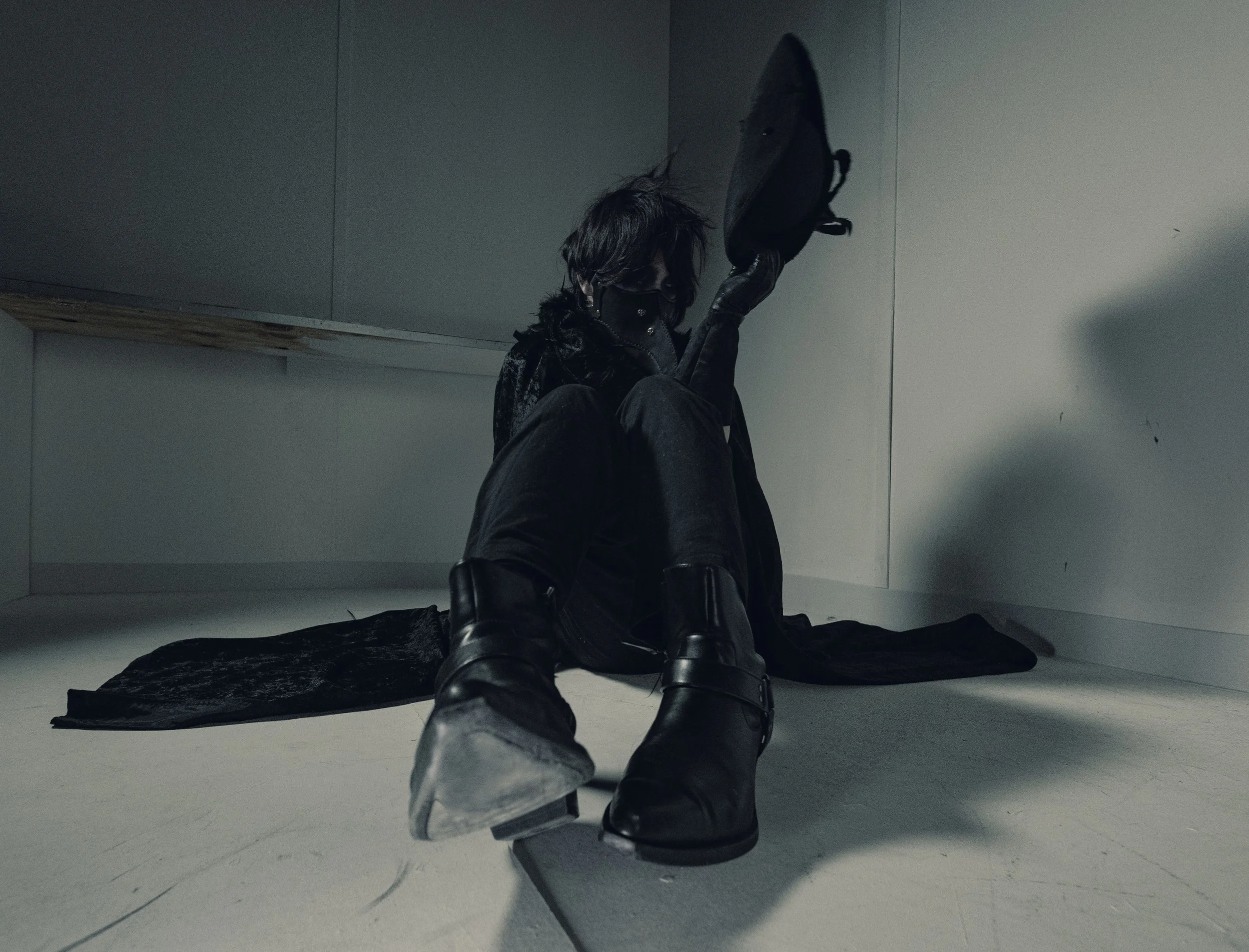 A person with dark hair sitting on the floor in a small, minimalistic room, wearing black boots and dark clothing, with a backpack leaning against the wall behind them.
