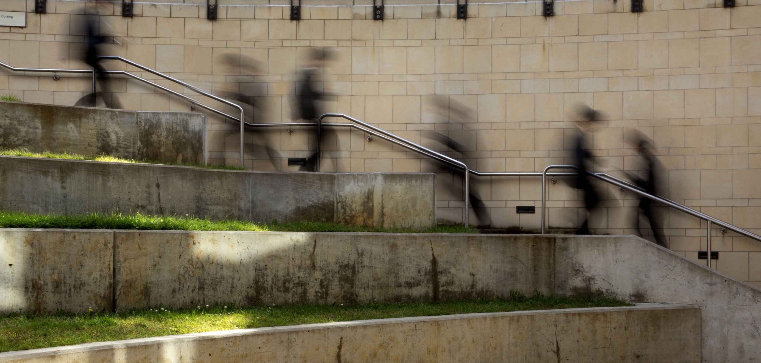People walking down an outdoor staircase with metal handrails, blurred motion, against a beige brick wall.
