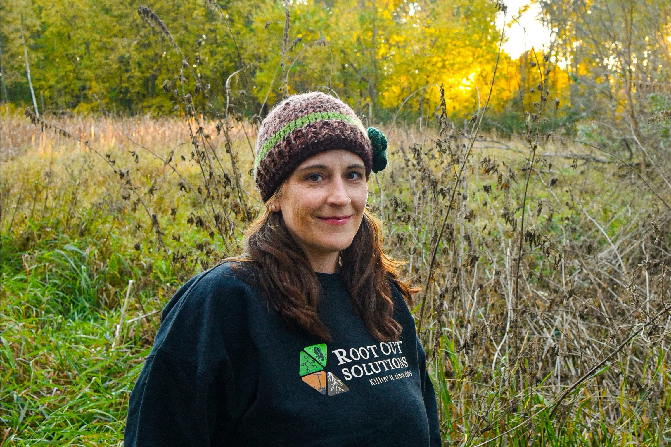 A woman with long brown hair, wearing a colorful knit beanie and a black T-shirt with a logo that reads 'Root Out Solutions,' stands outdoors in a field of tall grass and weeds during autumn, with trees and a setting sun in the background.