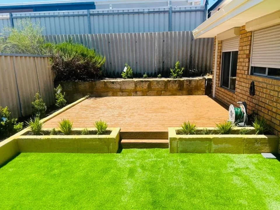 A backyard with a vibrant green lawn, decorative plants in raised brick garden beds, a wooden patio, and a brick house wall.