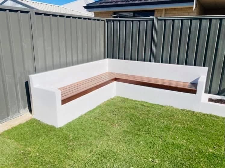 Outdoor corner seating area with a white concrete base and brown wooden bench in a backyard with grass and a metal fence.