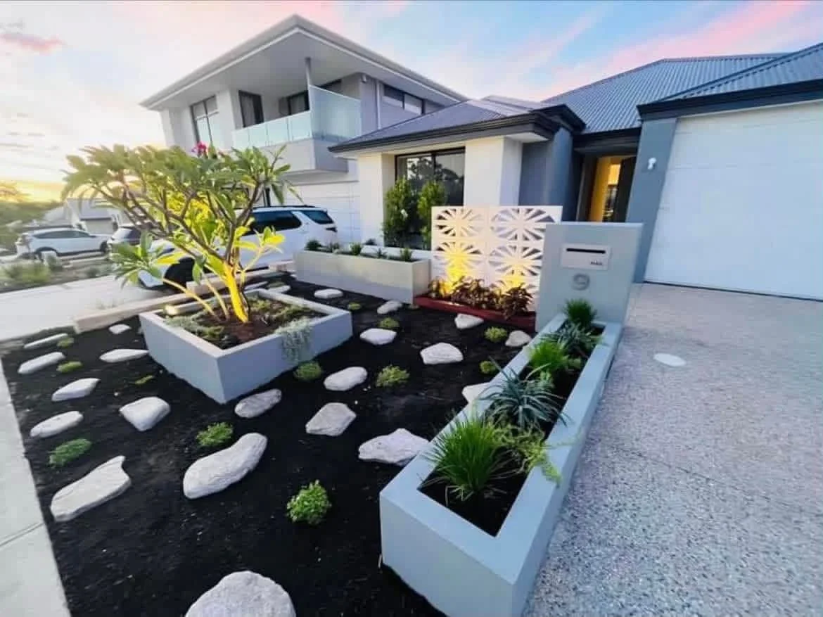 Modern house exterior with a landscaped front yard, featuring raised garden beds with plants, a tree, rocks, and pathway, during sunset.