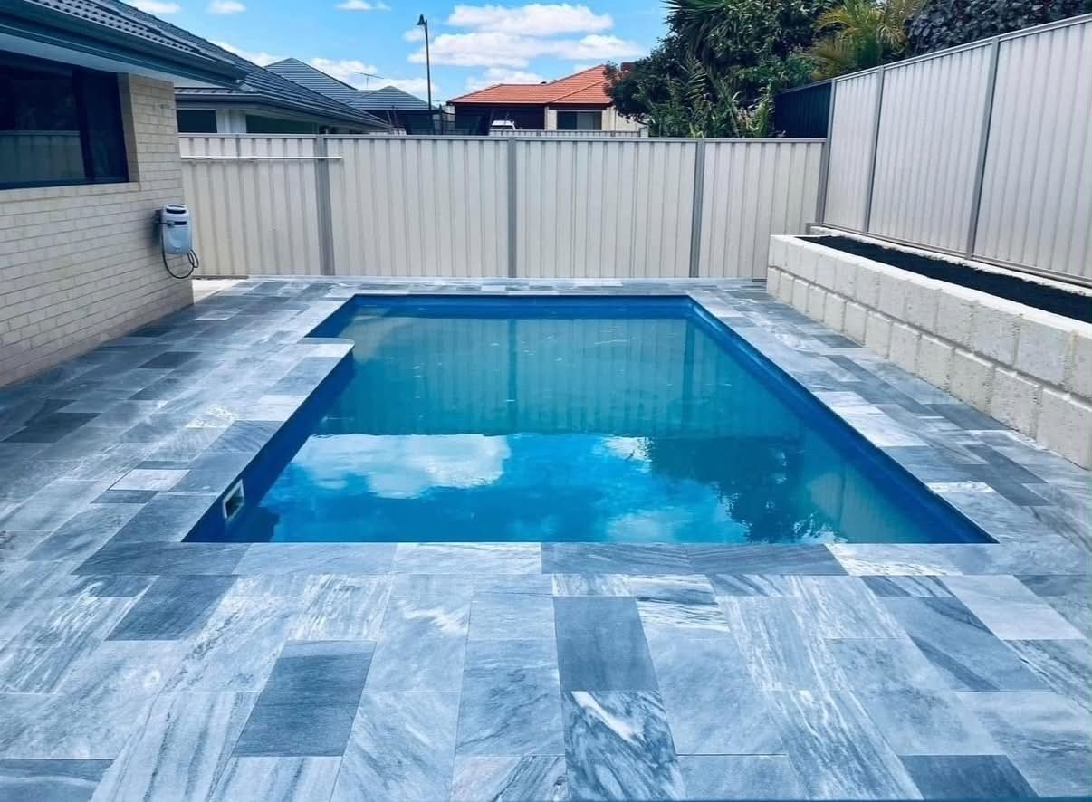 Backyard pool with blue water, surrounded by gray marble tiles, enclosed by beige fences, with neighboring houses visible in the background under a partly cloudy sky.