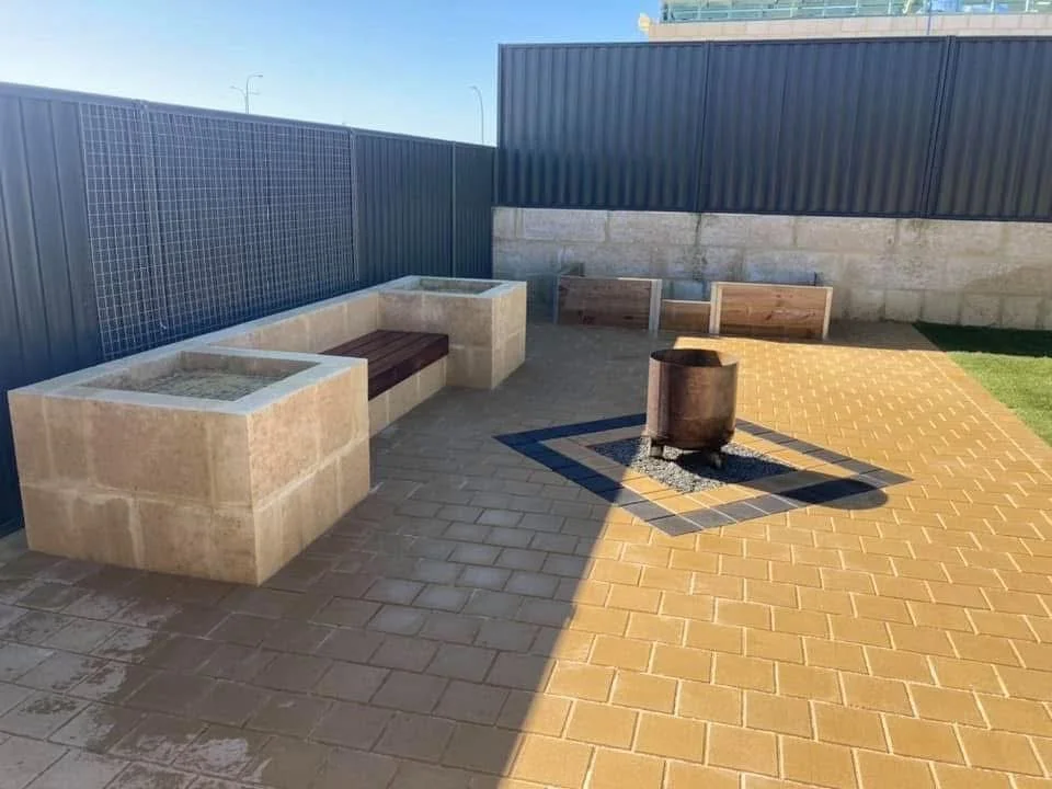 Backyard with concrete planters, a fire pit on a brick patio, surrounded by a wood and metal fence, with a small patch of grass.
