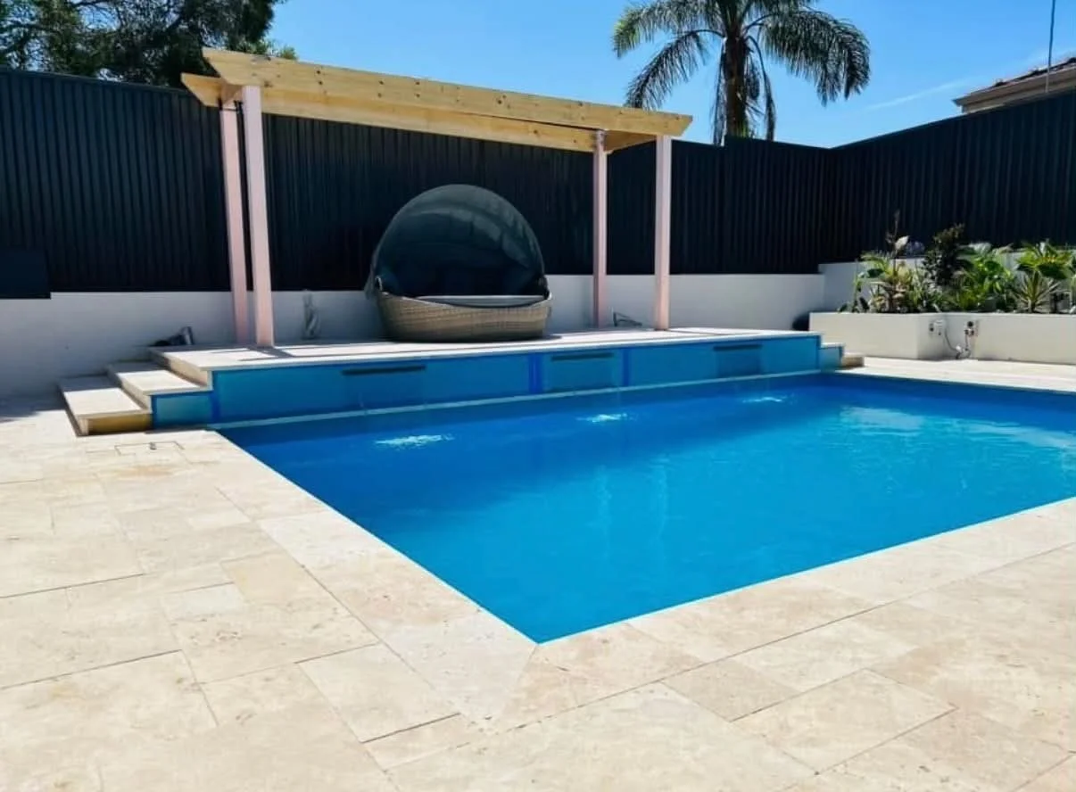 A backyard swimming pool with steps leading into the water, a sitting area with a large round chair under a wooden pergola, surrounded by a black fence and lush green plants, under a clear blue sky.