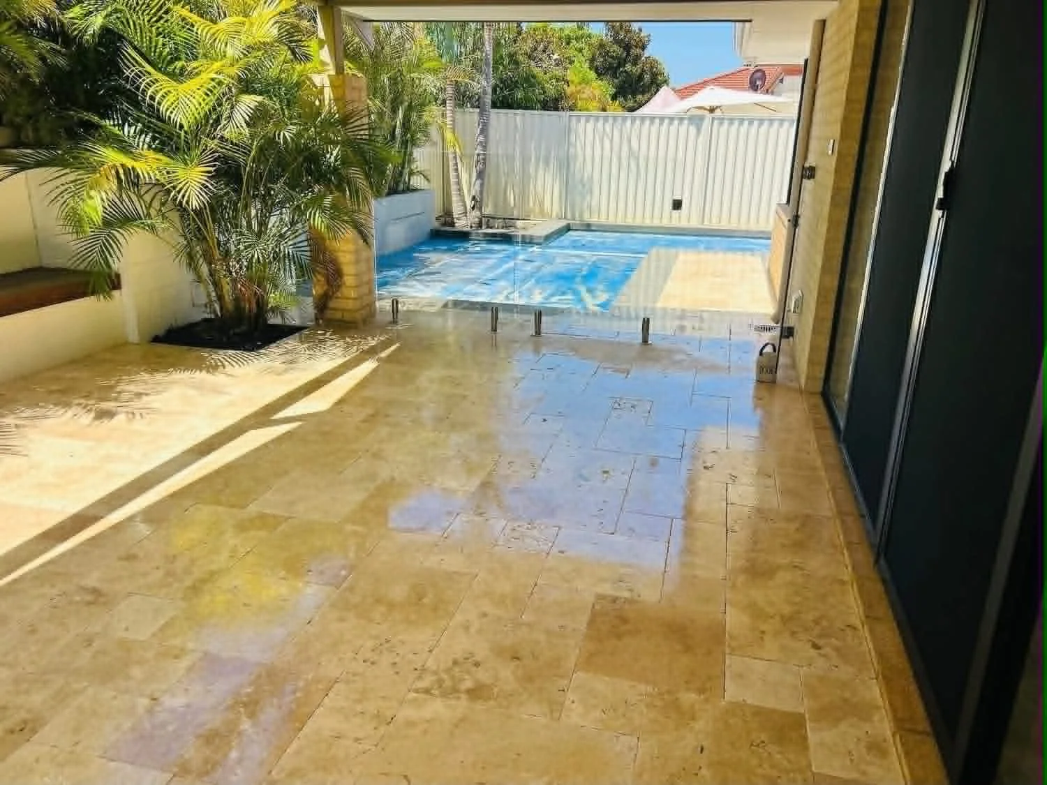 An outdoor patio area with beige tiled flooring, a large potted palm tree, a small pool with a blue liner, and a white fence in the background under a partly sunny sky.