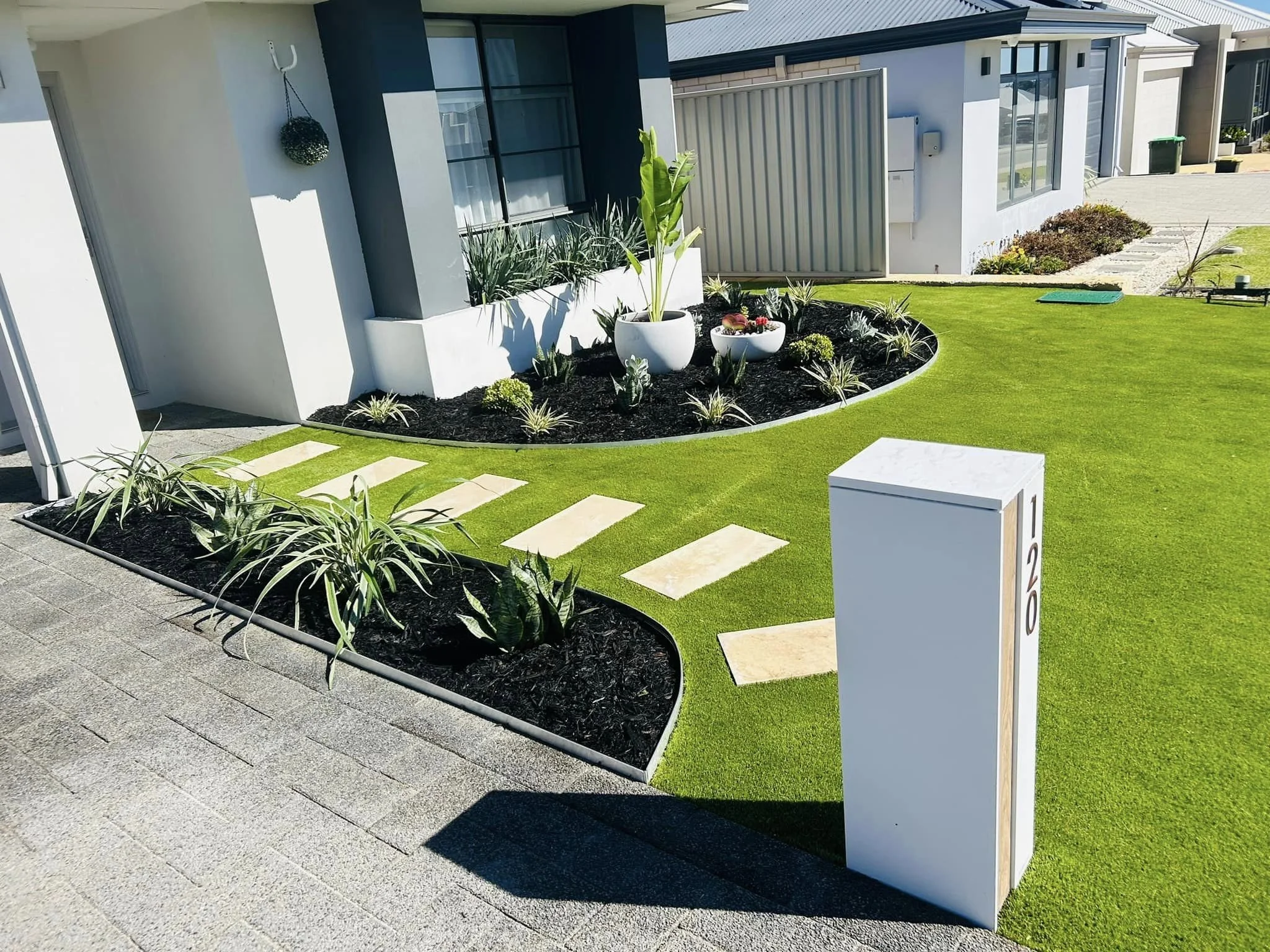 Front yard garden with small white planter pots containing green plants, bordered by a curved bed of dark mulch and green grass, in front of a modern house with large windows and a white mailbox.