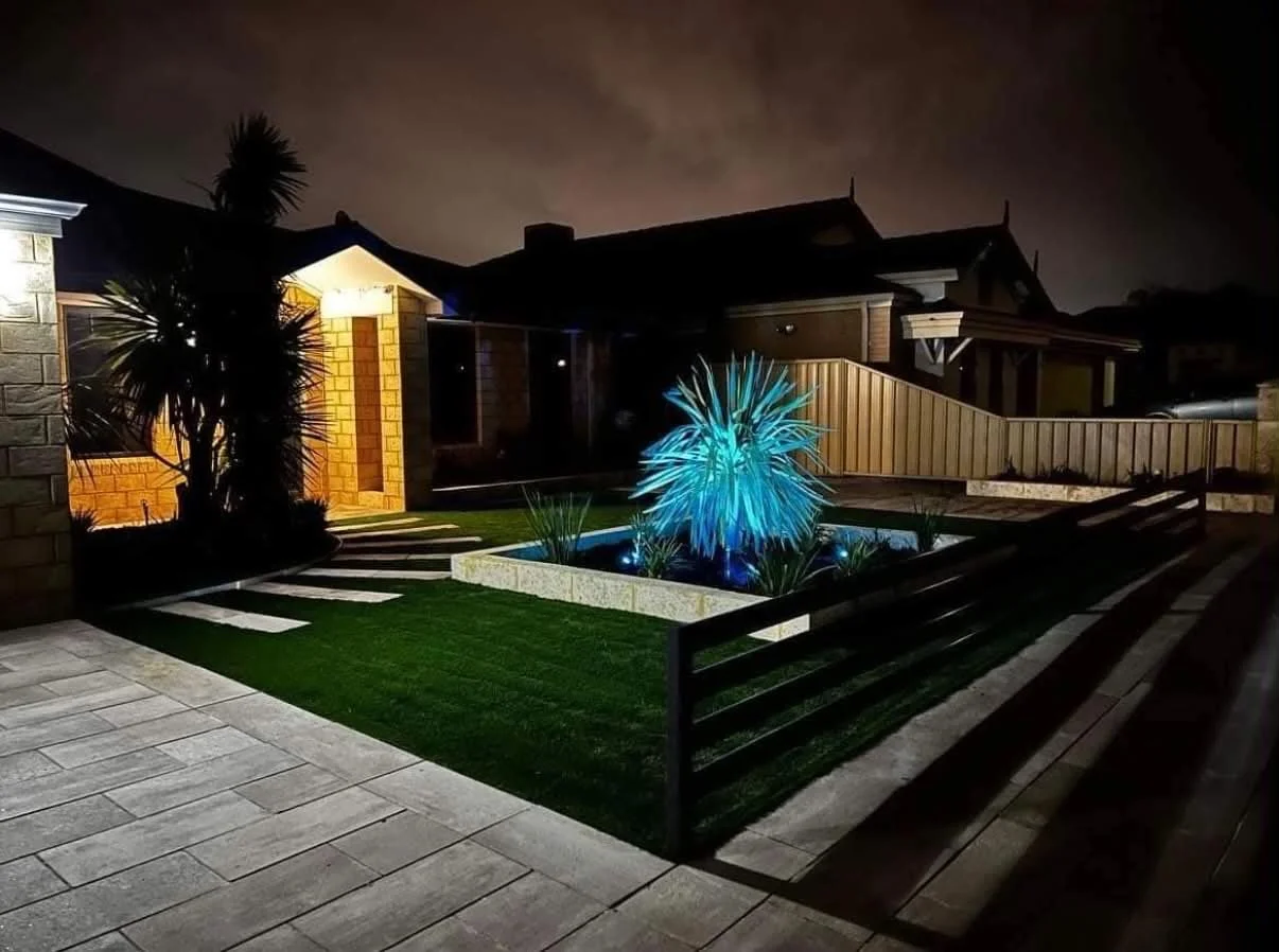 Night view of a modern backyard with a small water feature illuminated in blue, surrounded by plants and a lawn, with a brick pathway and a wooden fence.