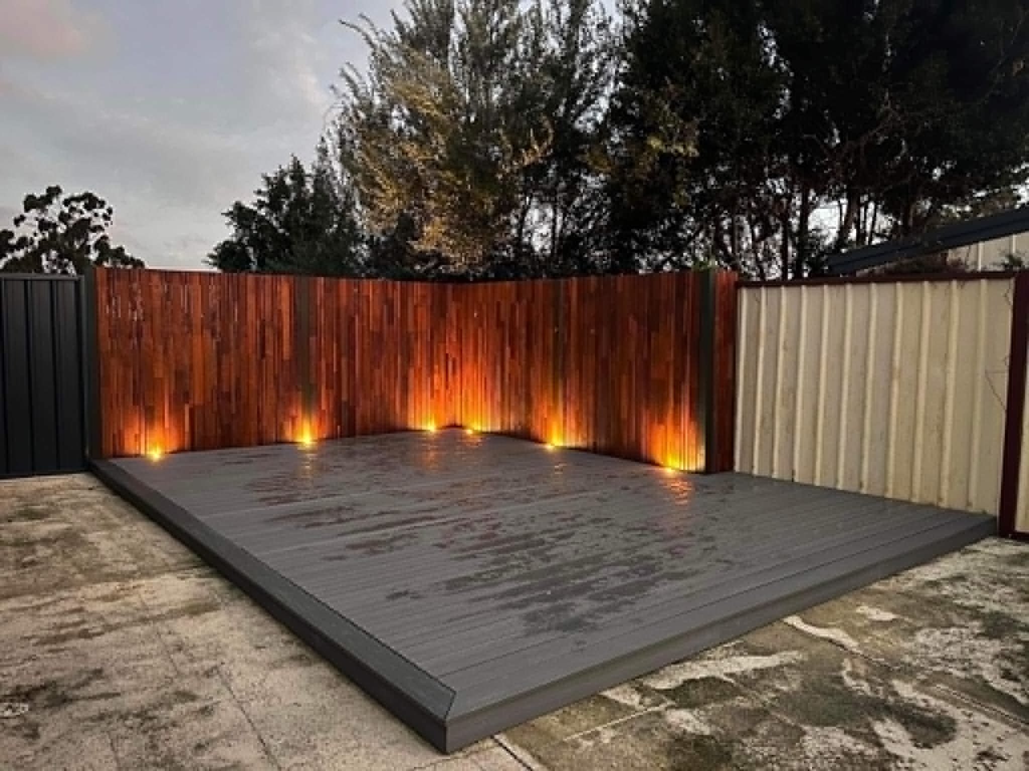 A backyard patio with a wooden fence illuminated by ground lights, a small deck, and trees in the background during evening or dusk.