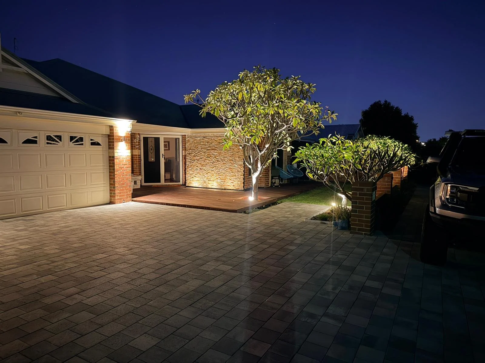 A modern house exterior at night with outdoor lighting highlighting trees and the entrance, a paved driveway, a garage, and parked vehicles.