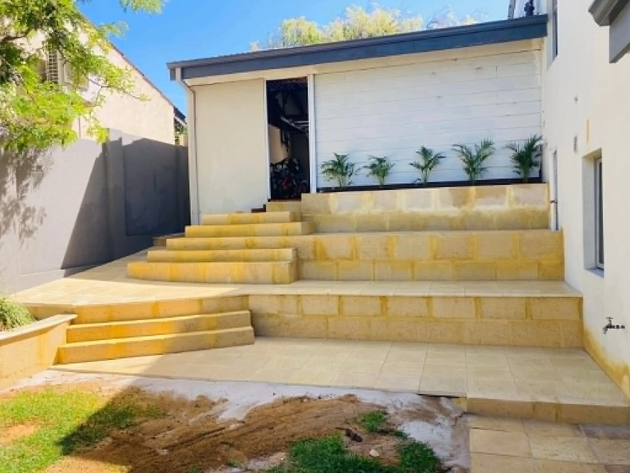 A backyard patio with stone steps leading up to a white house with a sliding glass door. There are small potted plants next to the door and a tree on the left side of the image. The sky is clear and blue.