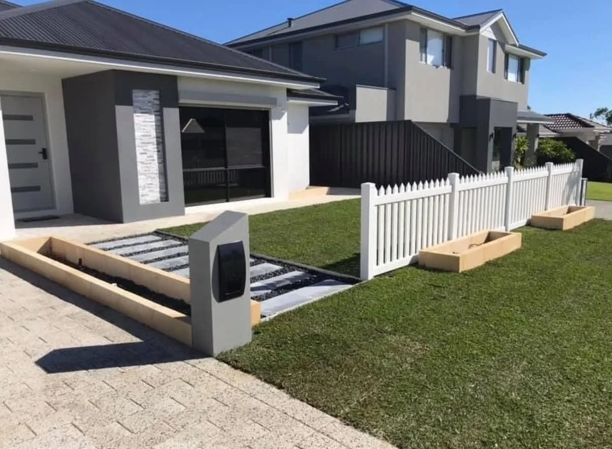 A modern house with a small front yard, white picket fence, planter boxes, and a mailbox. The house has gray and white exterior walls, a covered porch, and large windows. The yard features a grassy lawn and a paved walkway.