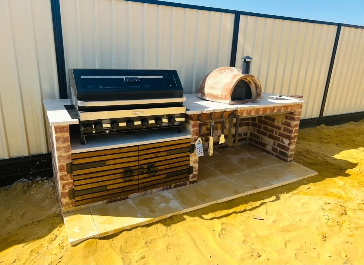 Outdoor brick and wood barbecue setup with a grill, a pizza oven, and hanging grilling tools, on a tiled surface with sand around.