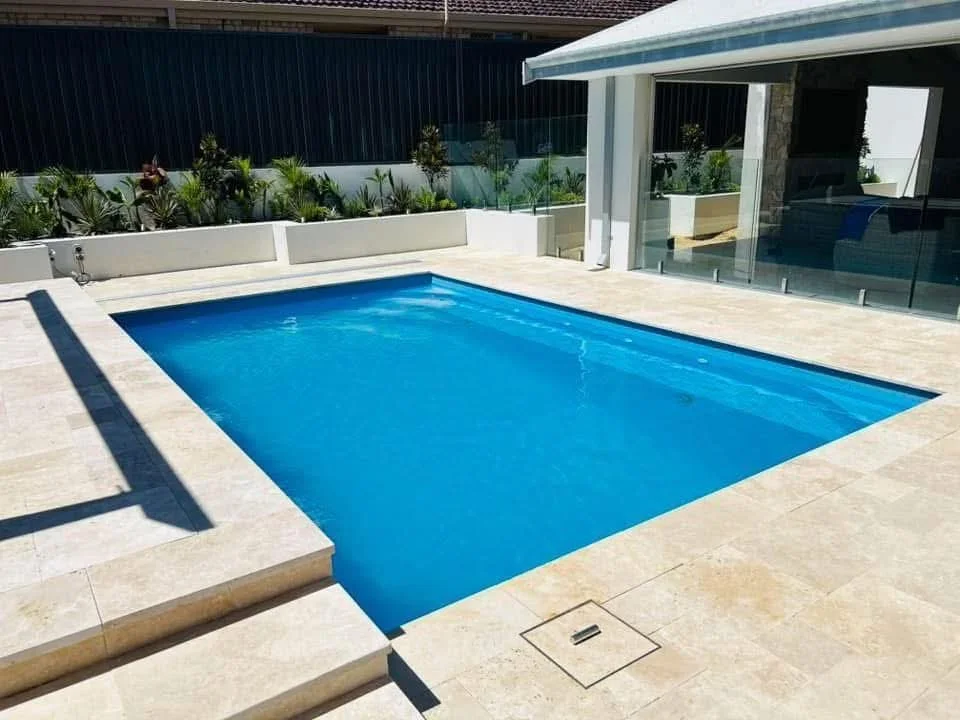 Backyard swimming pool with beige stone deck, glass railing, and modern house with large windows and white exterior.
