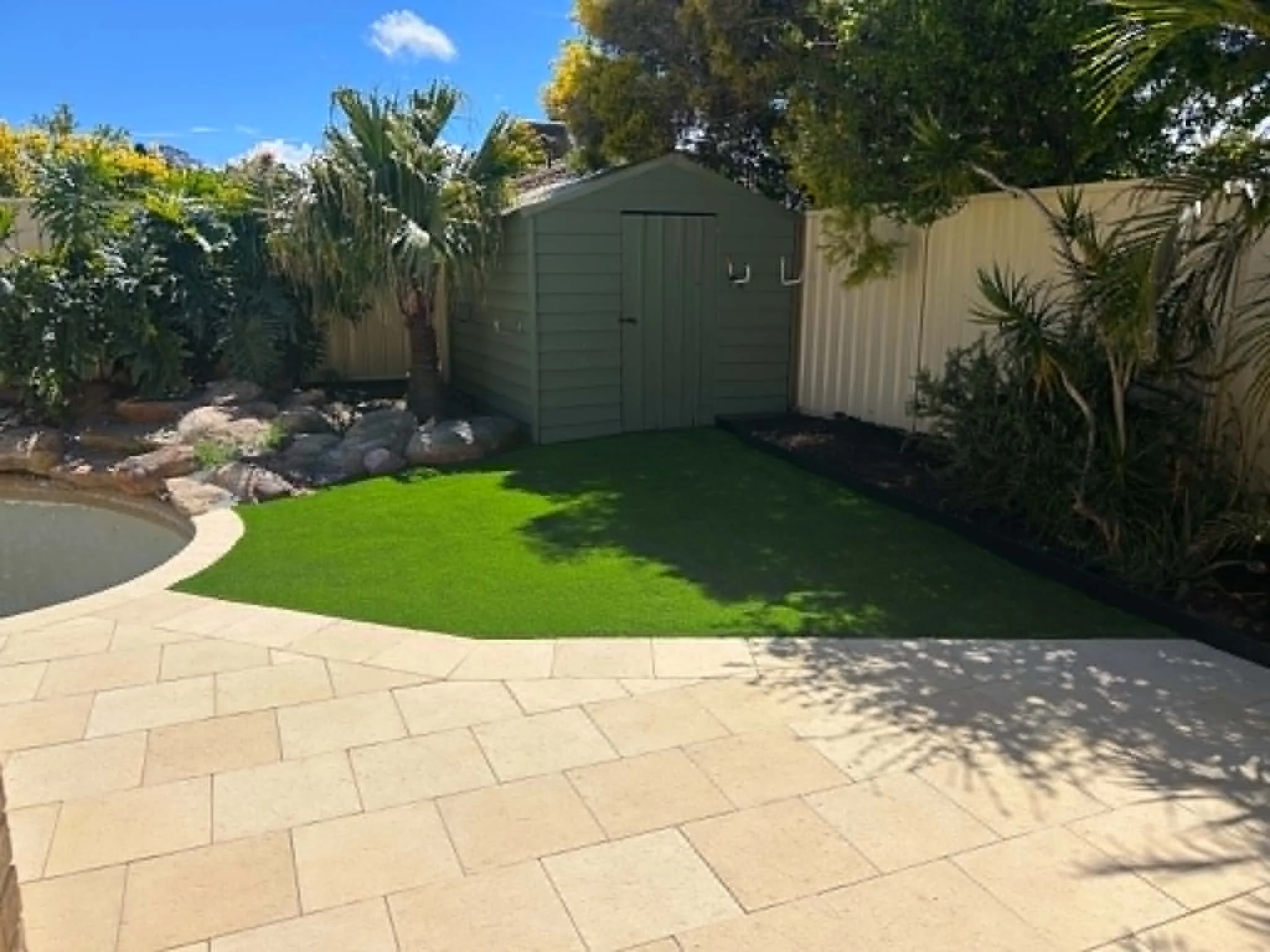 A backyard with a stone patio, a small lawn, a garden shed, and trees with some yellow flowers, under a blue sky with a few clouds.
