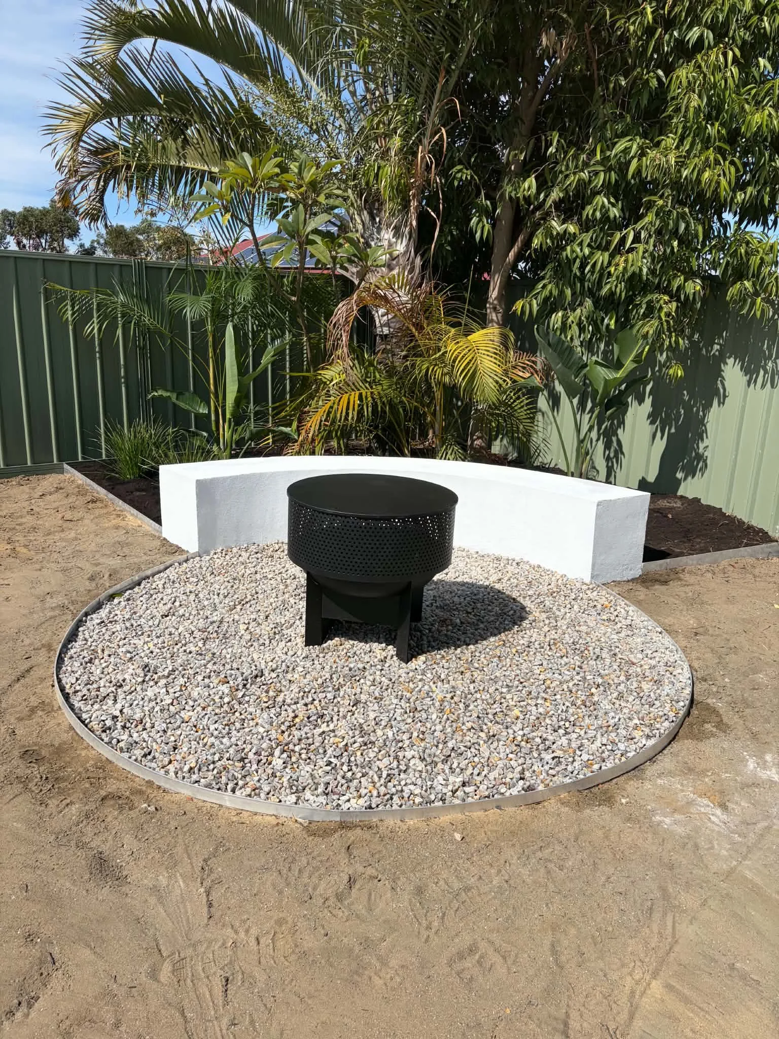 A backyard with a green metal fence, tropical plants, and a fire pit on a gravel circle. There is a white curved seating structure in front of the plants.