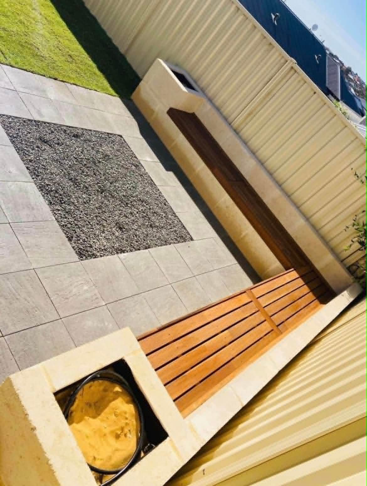 An outdoor patio area with tiled flooring, a wooden bench, a small table, and a wall-mounted wooden planter box. There is a yellowish wall with a black planter on a ledge and a green grass patch with a wooden fence and buildings in the background.