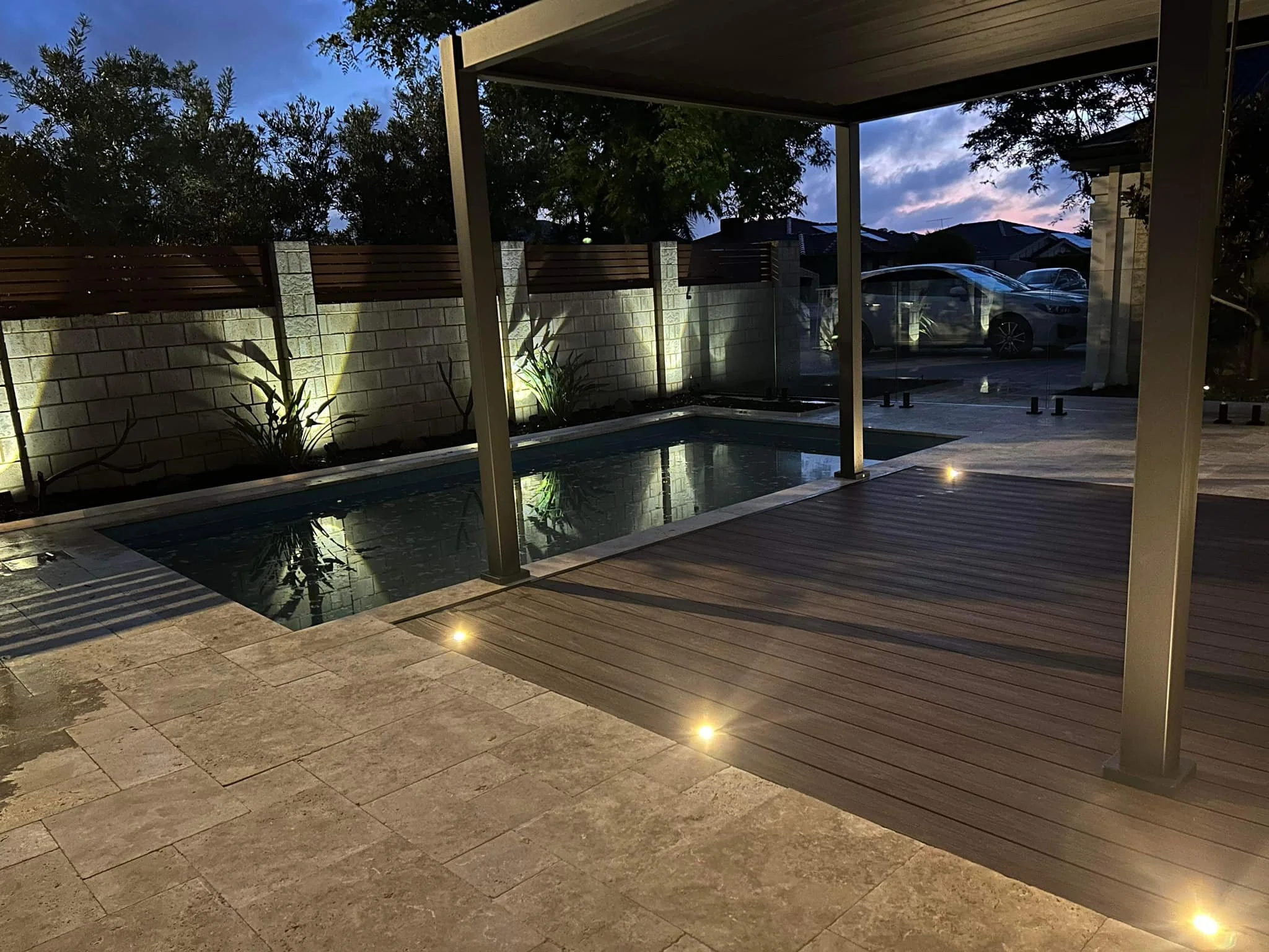 A backyard scene during dusk showing a swimming pool, outdoor lights, a stone patio, a wood deck, a wooden fence, trees, and parked cars in a driveway.