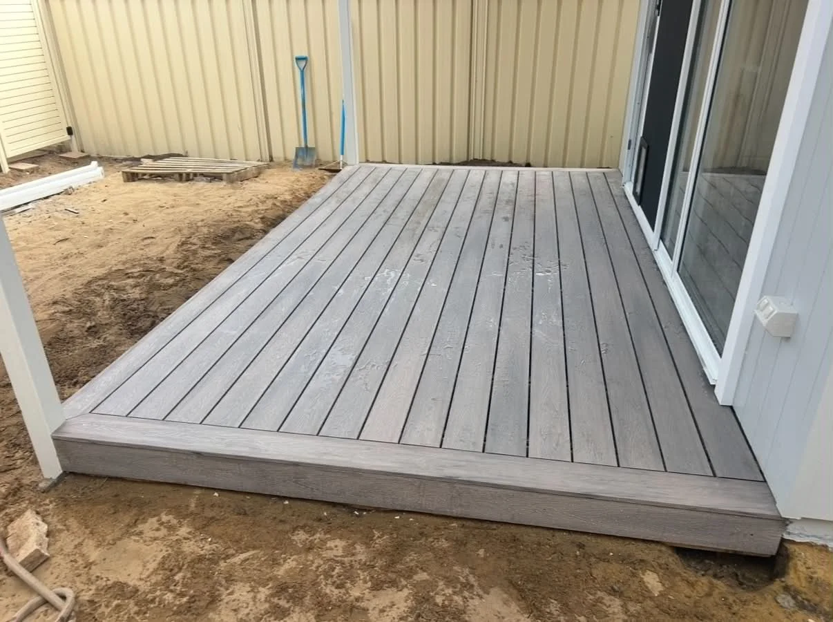 Newly installed wooden deck outside house with sliding glass door, surrounded by dirt and construction materials.