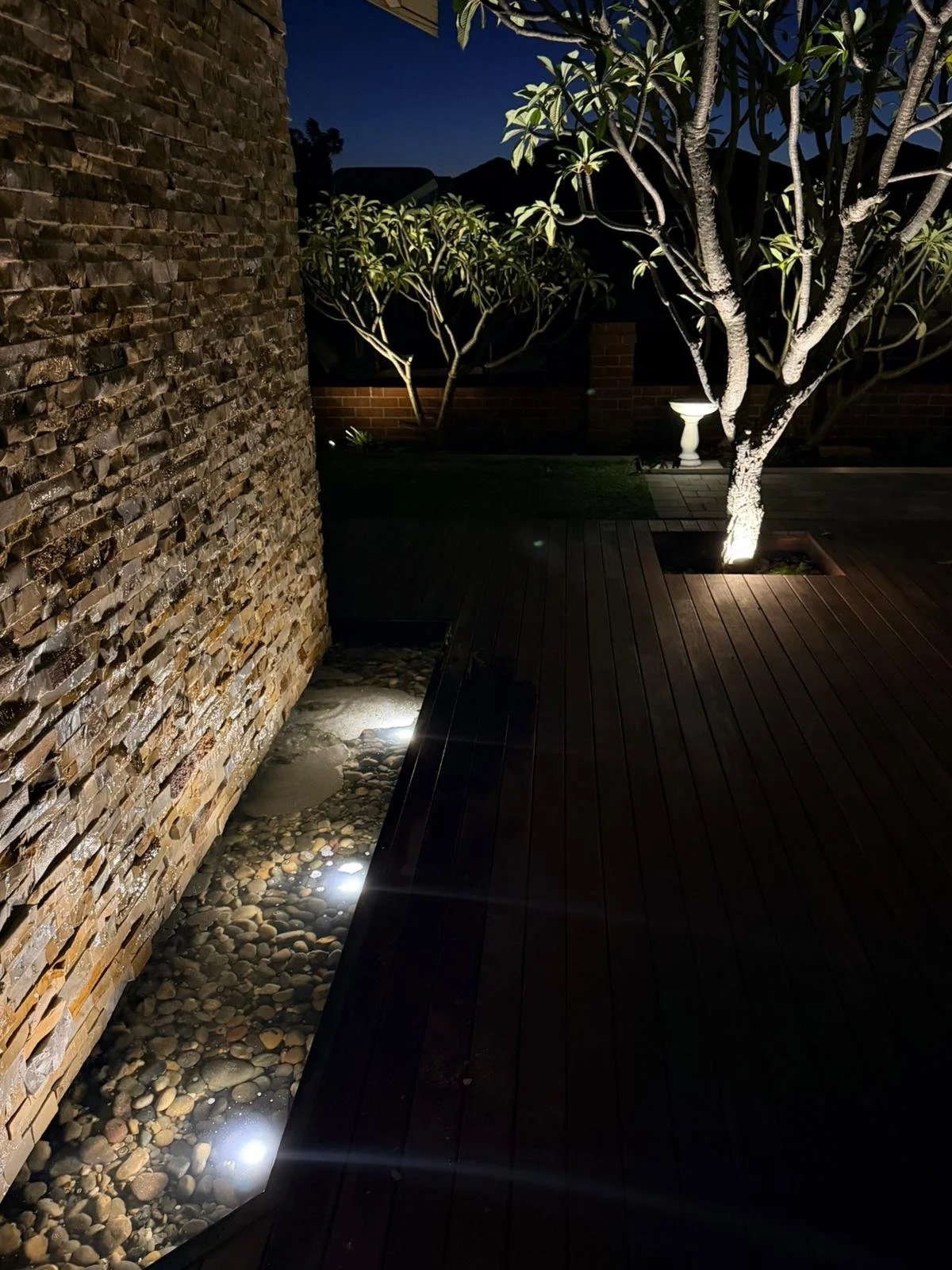 Nighttime outdoor patio with wooden flooring, illuminated trees, and a stone wall with ground-level lighting.