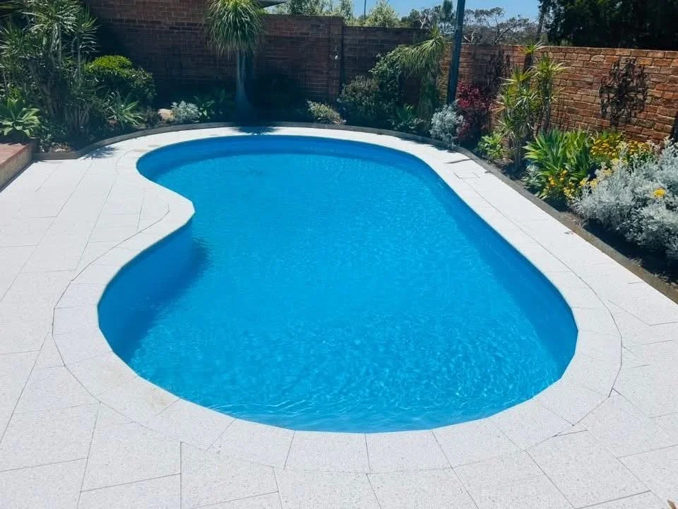 A backyard with a small kidney-shaped swimming pool, surrounded by white tiles and lush plants along a brick fence.