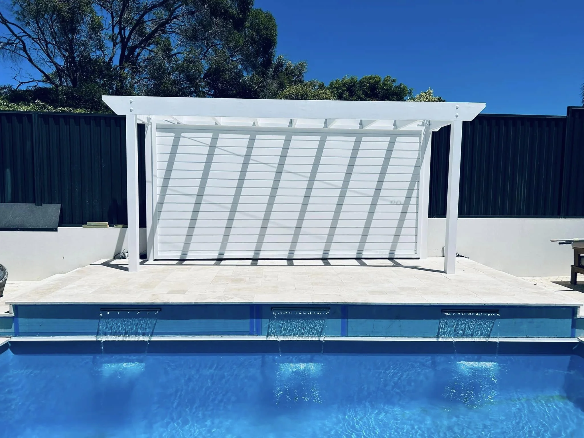 A modern outdoor swimming pool with blue water, a white trellis structure, and a black privacy fence. Tall trees with green foliage are visible behind the fence, and the sky is clear and blue.