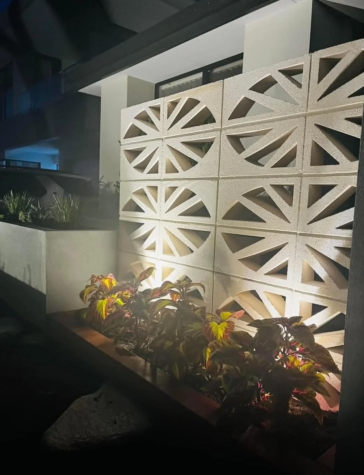 Nighttime exterior view of a modern building with decorative concrete block wall, illuminated from below, with plants in a planter in front.