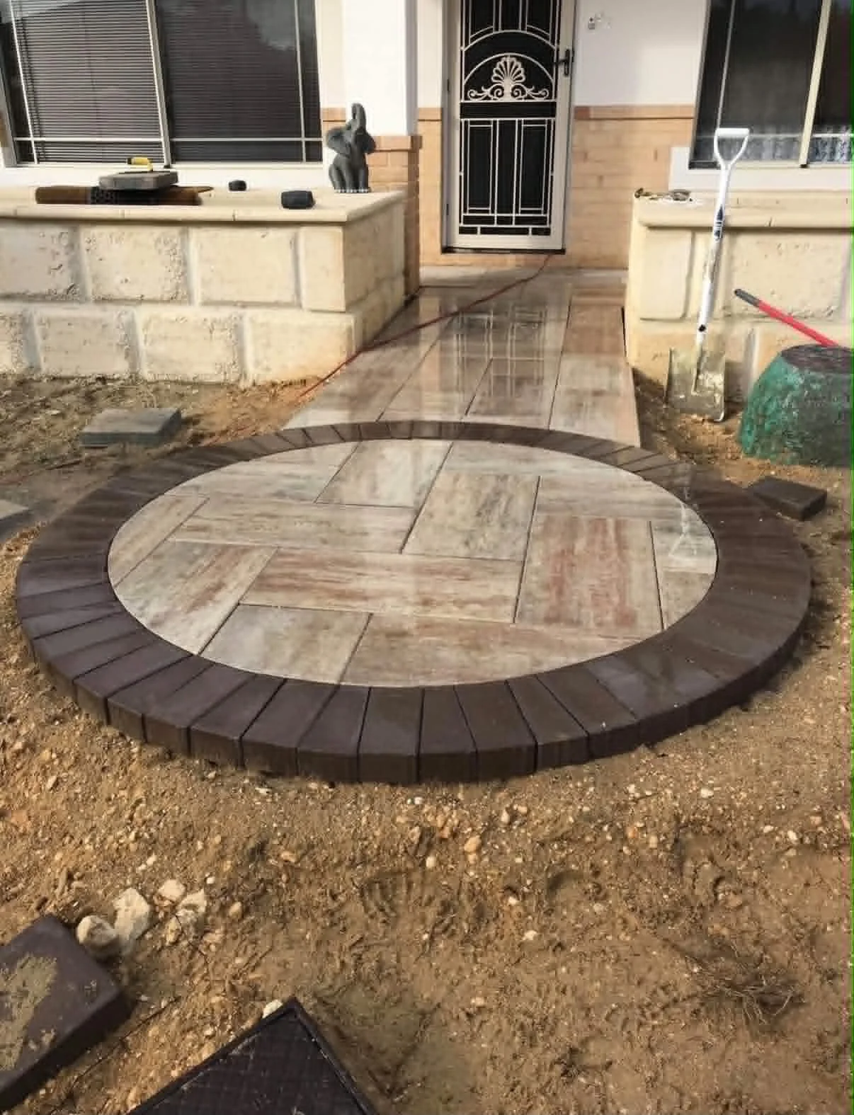 A circular tiled walkway in front of a house, with a brick border, partly surrounded by dirt and construction materials.