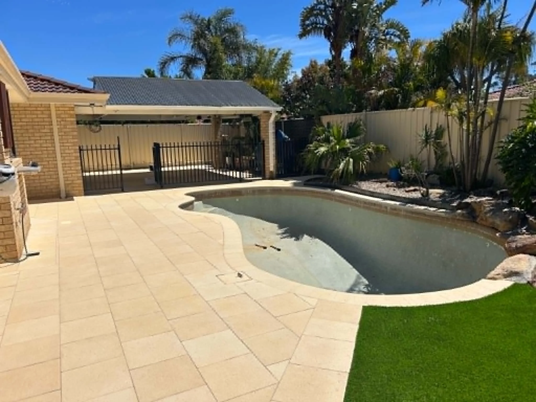 A backyard with a concrete patio, a small pool, a fenced area, and some greenery including trees and bushes under a blue sky.