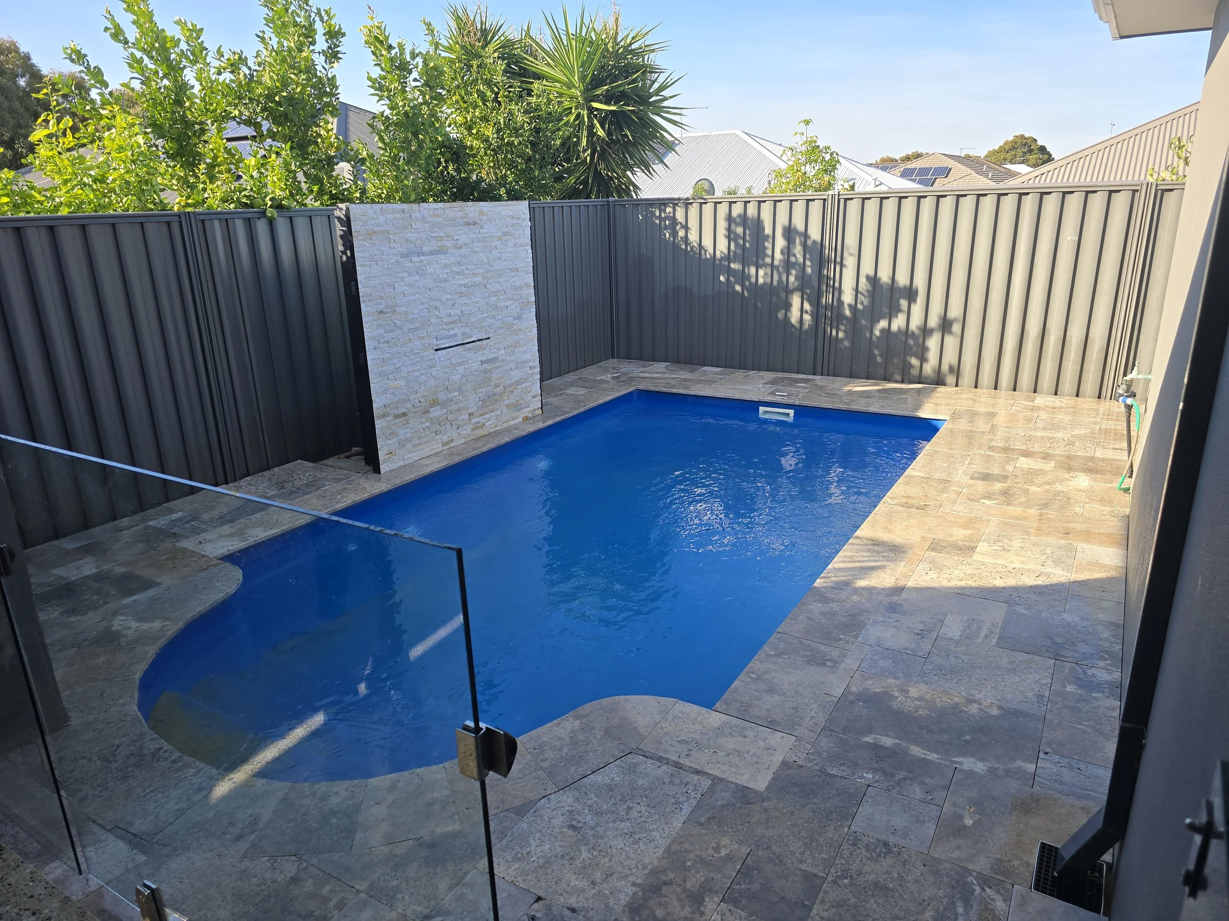 View of a backyard with a small swimming pool, surrounded by grey tiling and a tall black metal fence. There is a white stone feature wall with a narrow opening and built-in shelf, with lush green trees and neighboring houses visible in the backgroun