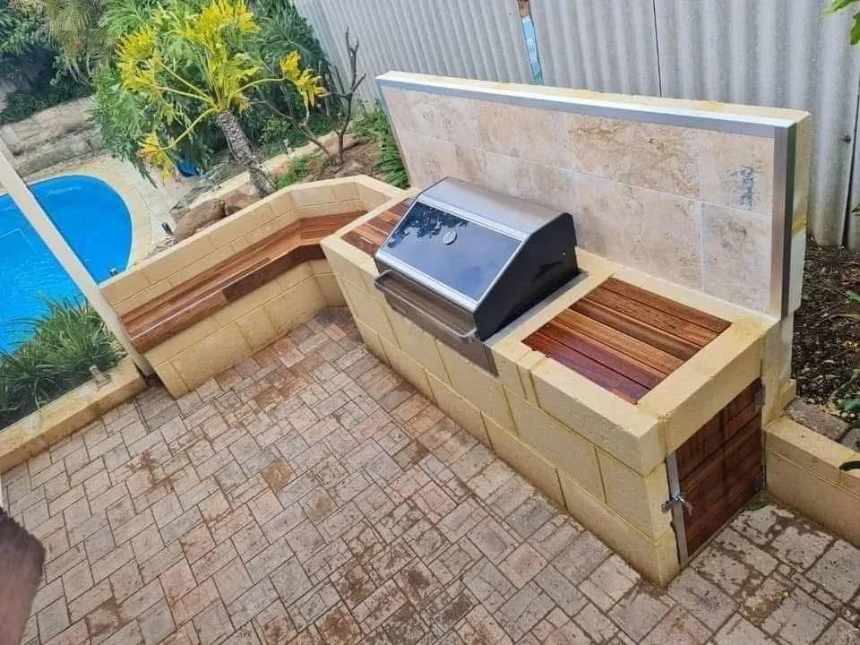 Outdoor brick and stone grill with a built-in lid and wooden panel, located in a backyard with a swimming pool and greenery.