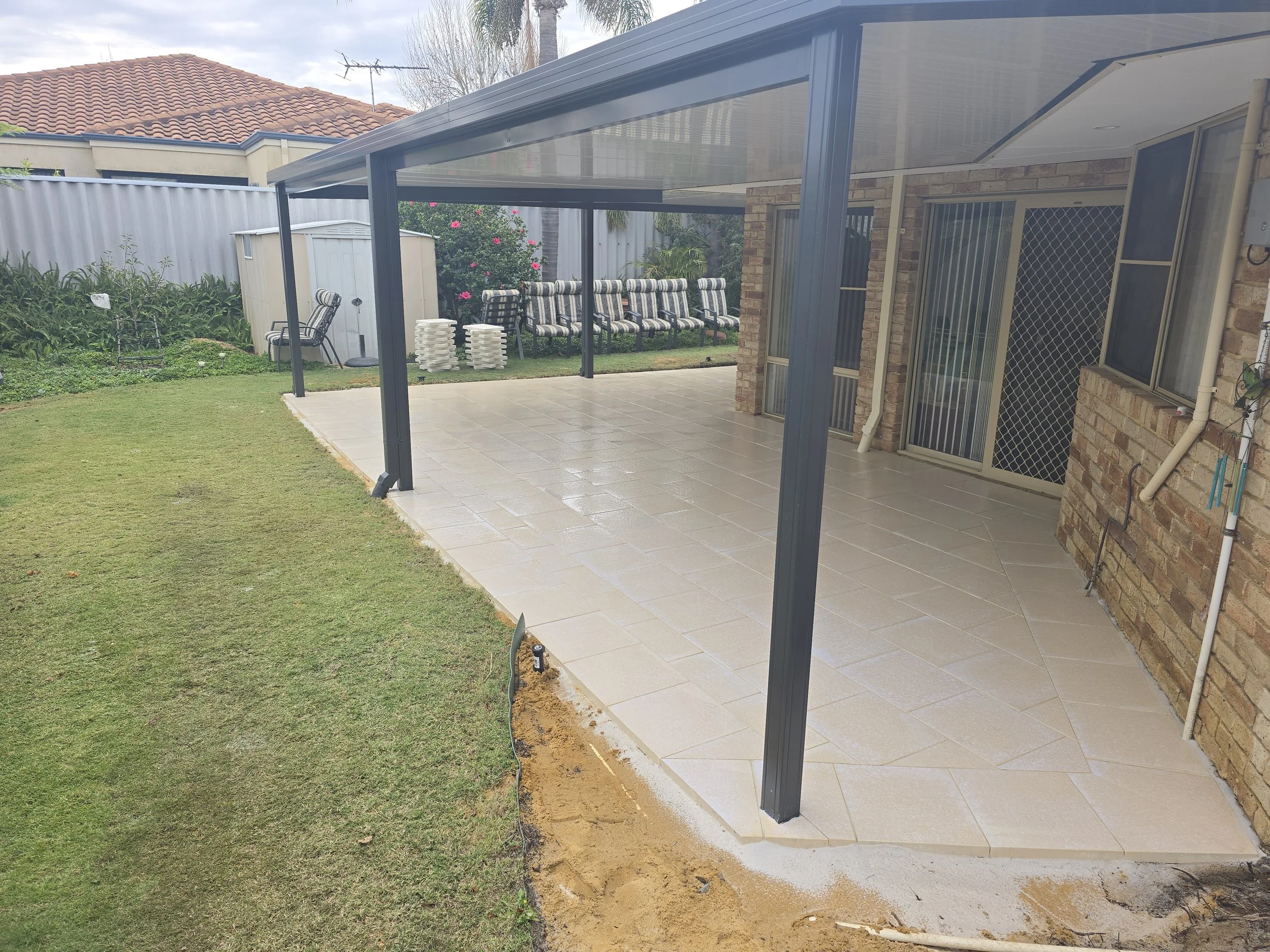 A backyard with a covered patio that has a metal frame and tiled flooring, adjacent to a grassy lawn. There are multiple striped patio chairs, a stack of white lattice panels, and a small shed. Part of a house with brick walls, windows, and a sliding