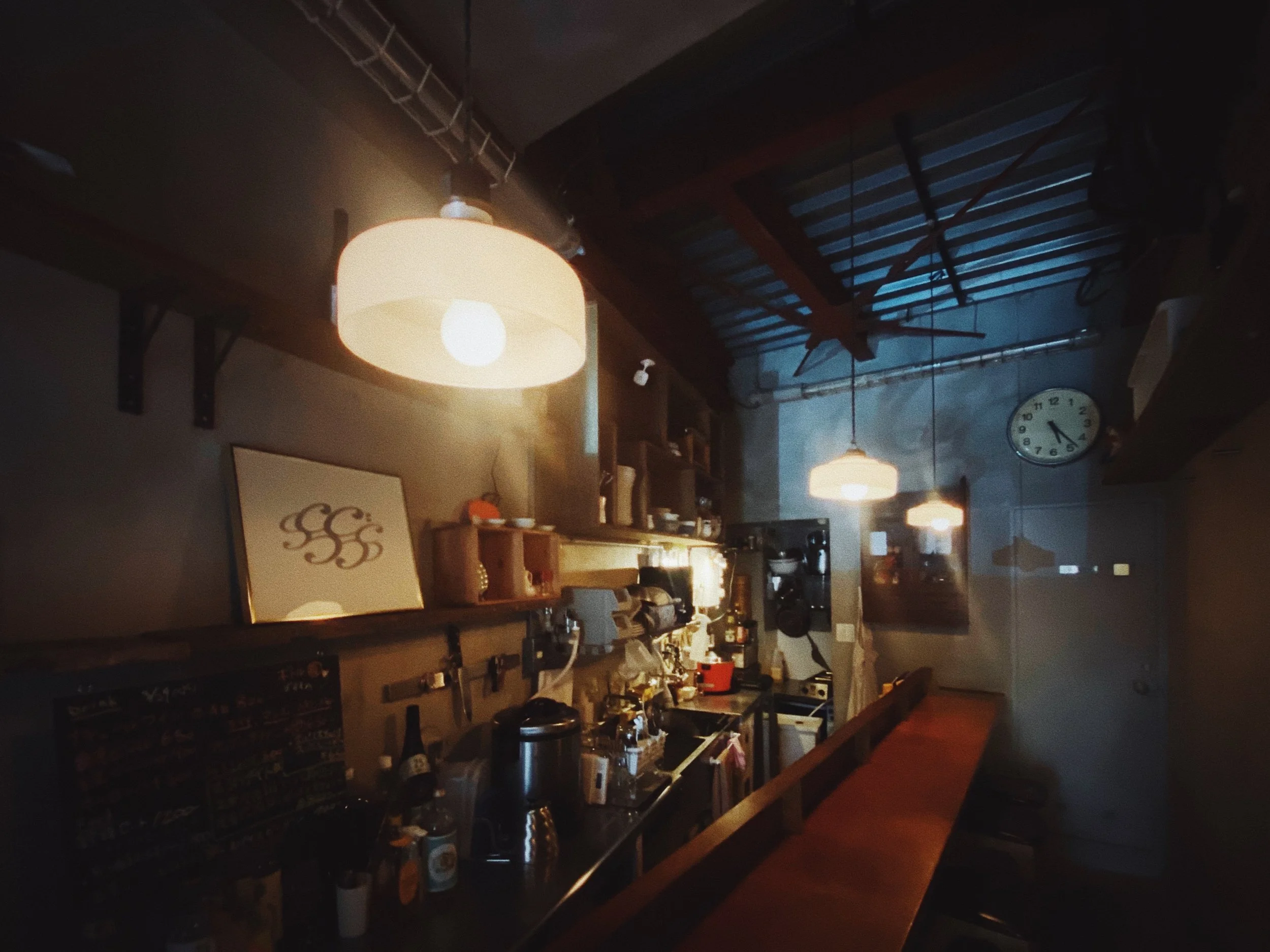 Inside view of a dimly lit café or restaurant with hanging pendant lights, a wall clock, and a bar area with various equipment and decorations.