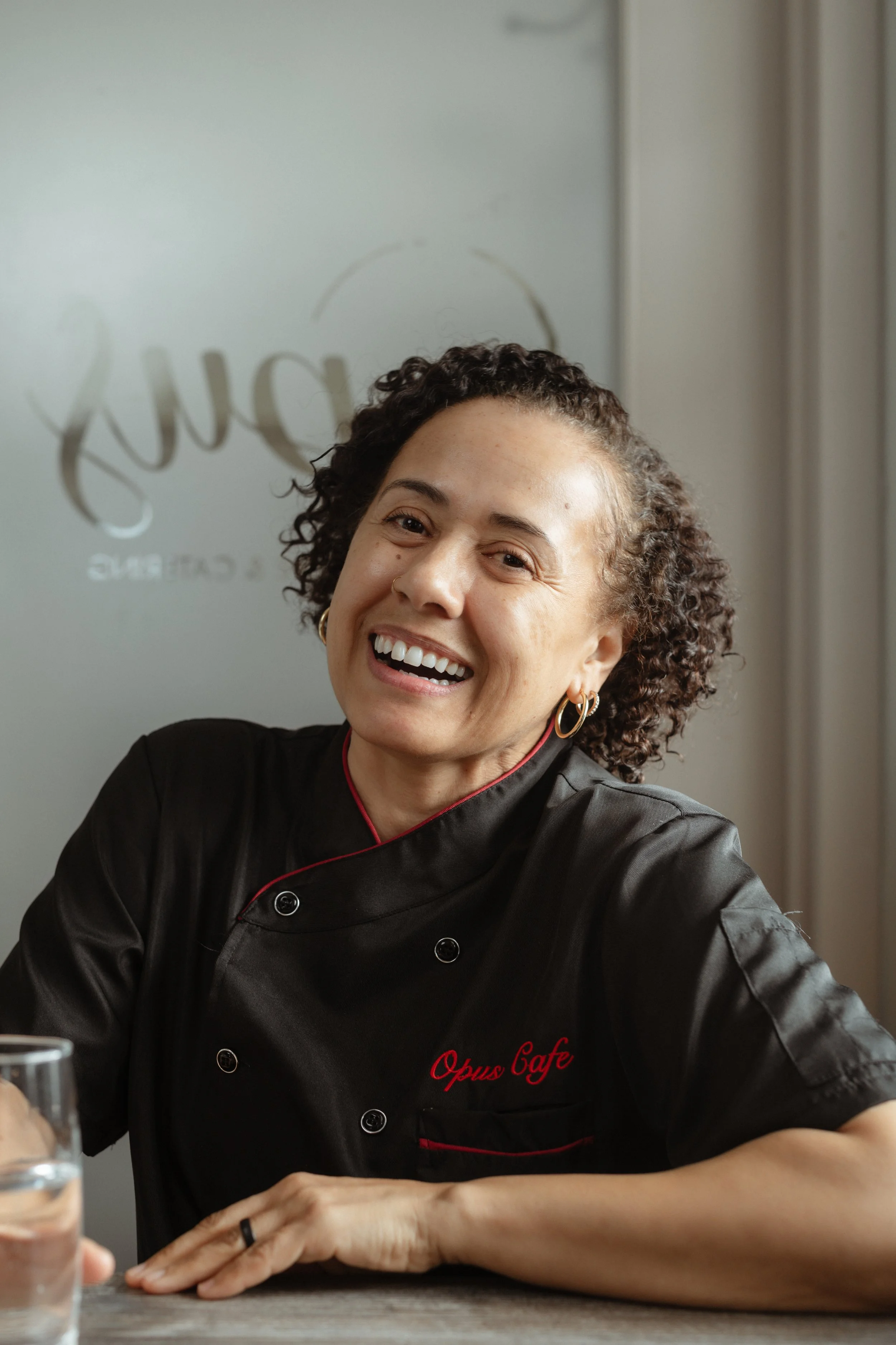 Opus Cafe Owner in a black chef jacket with red accents, with 'Opus Cafe' embroidered, sitting at a table inside a cafe.