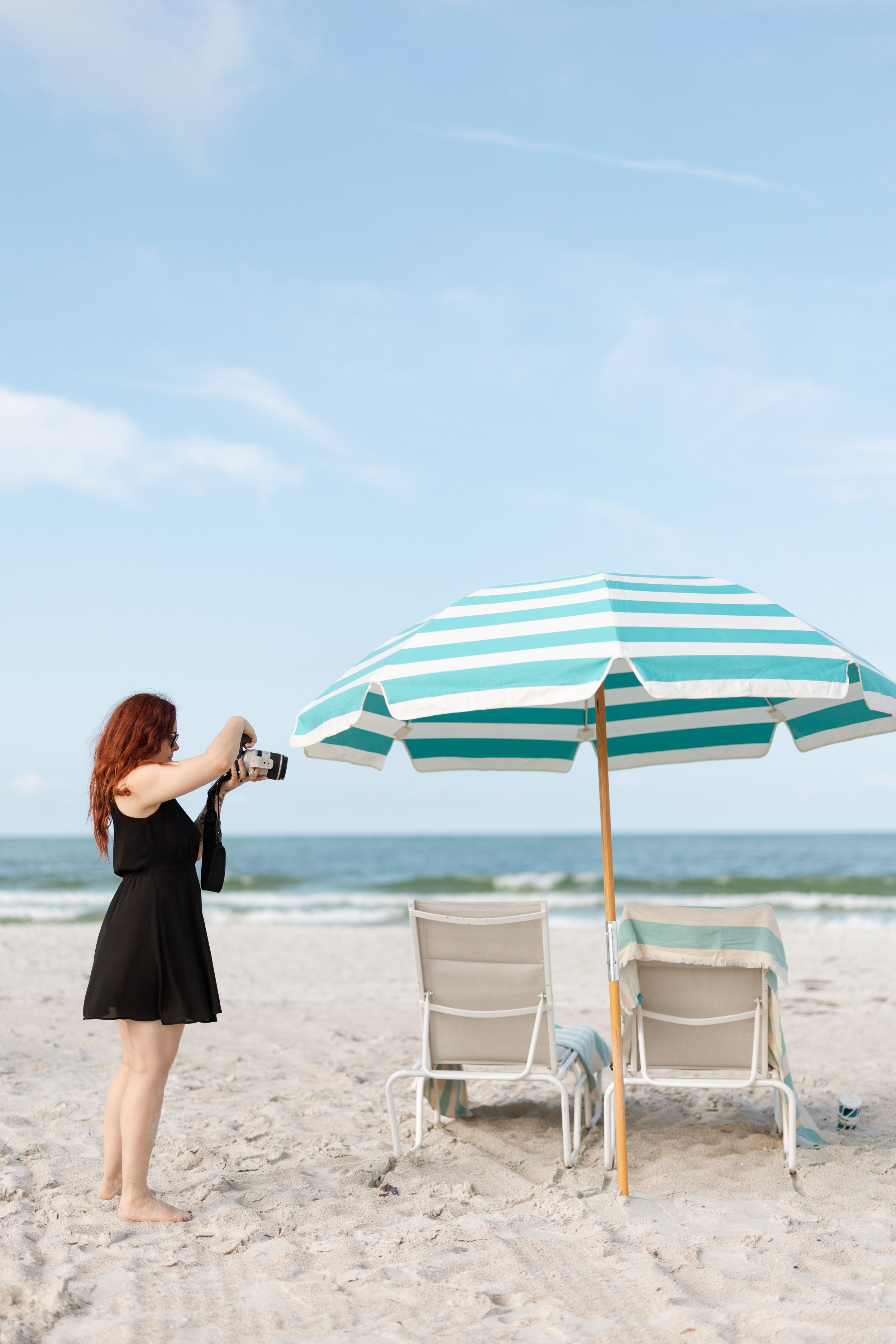 Woman with long red hair in a black dress taking a photo with a camera on a sandy beach with a blue and white striped umbrella, lounge chairs, and the ocean in the background.