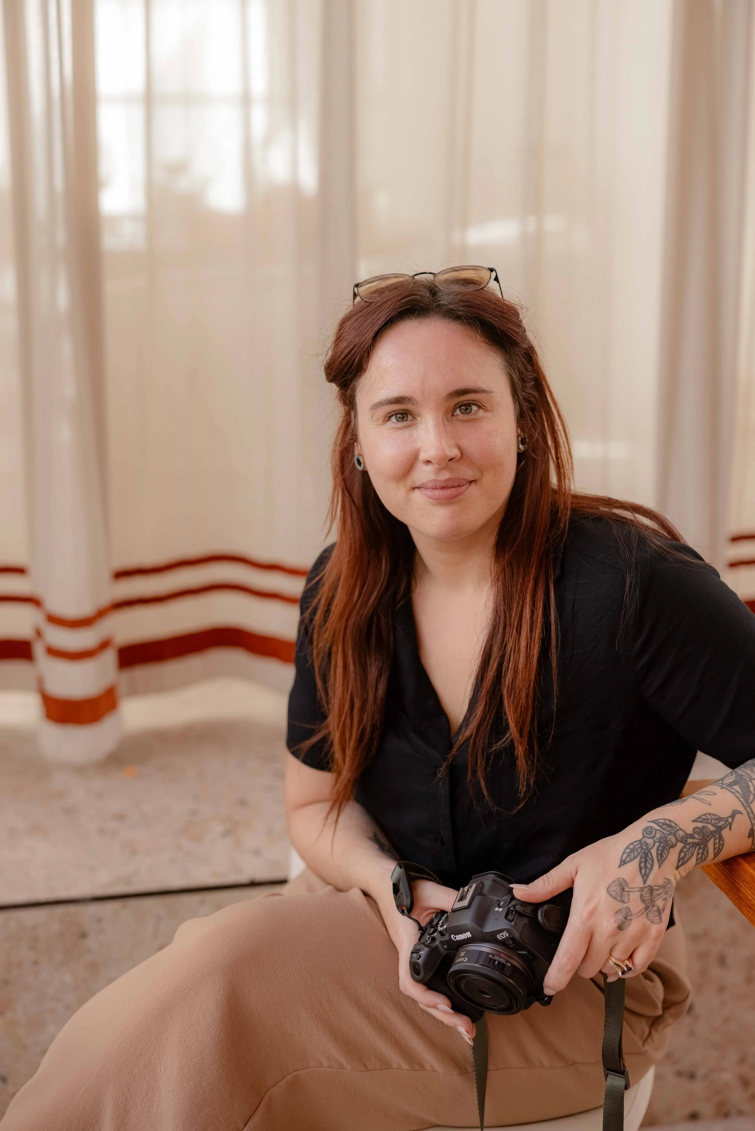 A woman with reddish-brown hair, glasses on her head, and tattoos on her arm, holding a DSLR camera, sitting indoors near cream-colored curtains with red stripes.